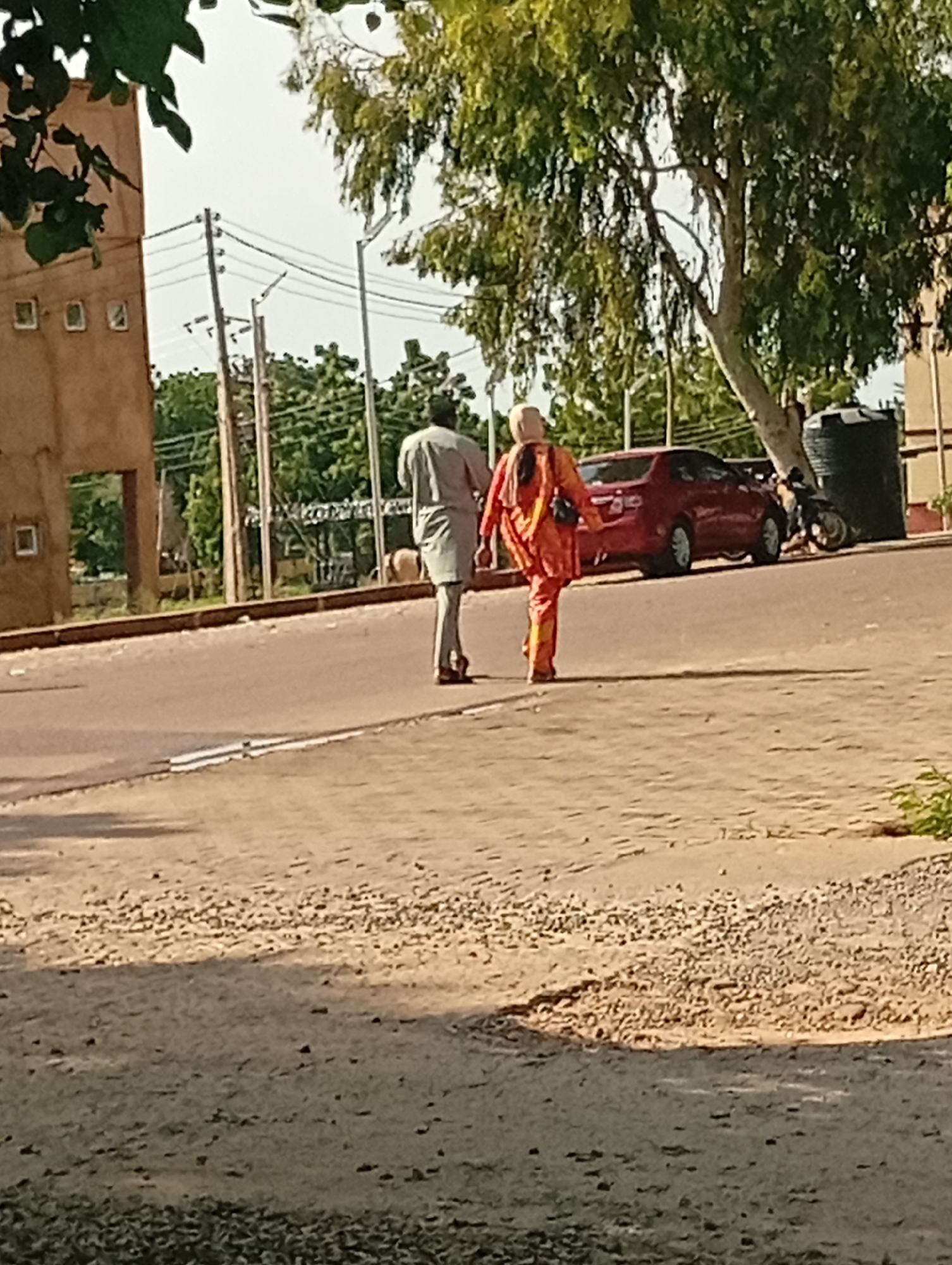 The image captures a street scene in Gombe, Nigeria. Two individuals, a man and a woman, are walking away from the camera on a paved road. The man is wearing a grey traditional outfit. The woman is dressed in a bright orange outfit, with matching trousers and a tunic. Behind them, a red car is parked on the side of the road, and a black motorcycle is visible next to a large black water tank. The background features lush green trees, utility poles with electrical wires, and the facade of a building. The ground in the foreground is a mix of sand and gravel. The lighting suggests it is daytime, possibly late afternoon due to the shadows.