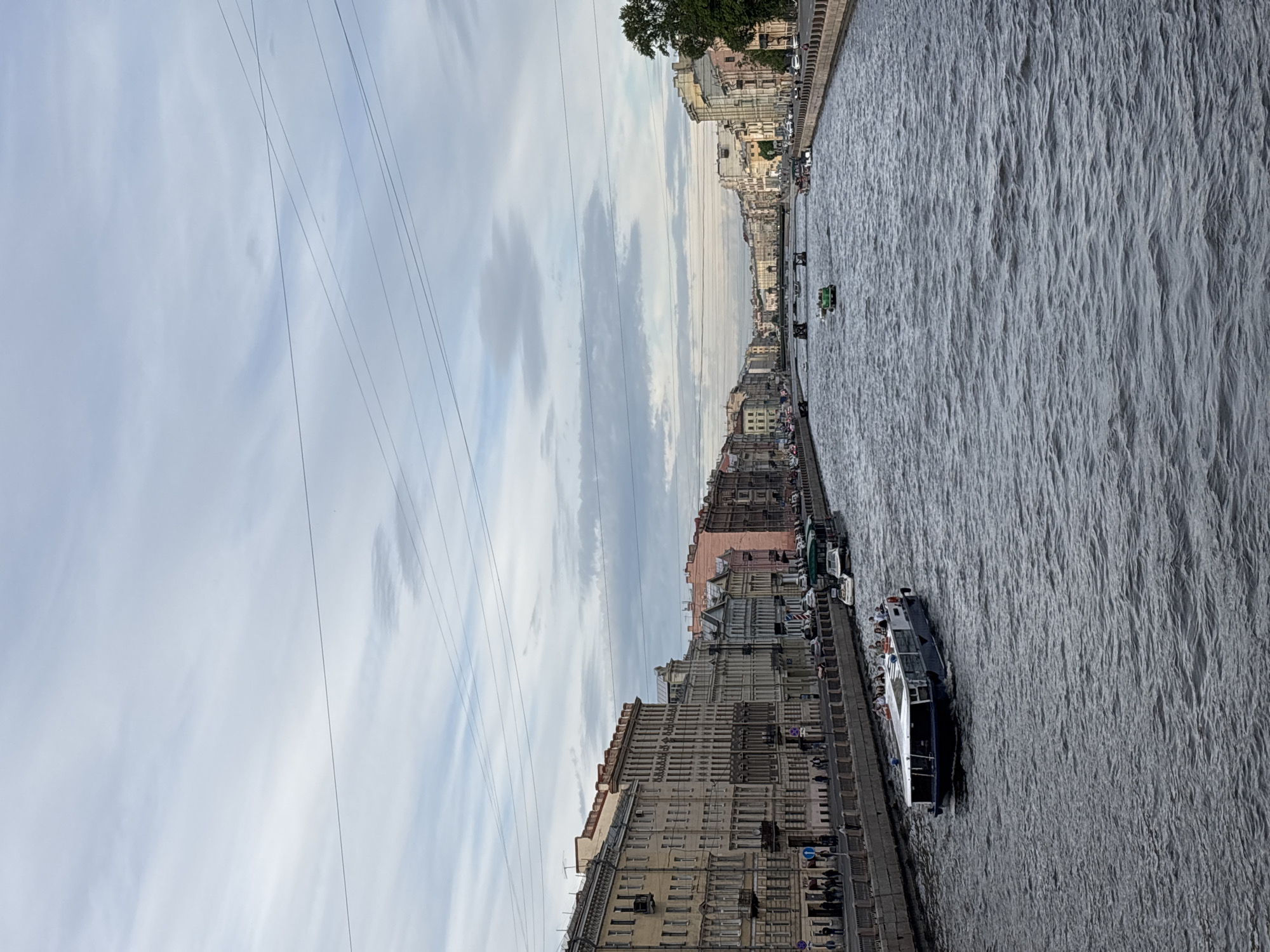 The image captures a panoramic view along a wide canal or river in Saint Petersburg, Russia, under an overcast sky. The scene is dominated by the waterway in the foreground and center, with historic city buildings lining both banks.
On the left bank (after mentally rotating the image), a long stone embankment features railings and streetlights, with small figures of people walking along it. Several blue circular signs with white arrows, indicative of traffic or pedestrian regulations, are visible. One clearly shows a white arrow pointing forward. Alongside the embankment, a large passenger boat with a dark hull and lighter upper deck is either docked or moving slowly, carrying passengers who are visible on its open deck. Further down the canal, another smaller green-hulled boat is also present.
The banks are densely populated with multi-story buildings, characteristic of 18th- and 19th-century European architecture, in muted tones of beige, grey, and some with soft pink or orange hues. Windows are numerous, and the buildings recede into the distance, suggesting a long stretch of urban development. A few trees with green foliage are visible on the far bank towards the horizon.
The sky is largely cloudy, with a mix of light grey and white clouds, suggesting an overcast day, possibly late morning or afternoon. Several thin, dark lines, likely power or tram wires, crisscross the upper sky. The surface of the water is choppy and textured, reflecting the grey sky and creating ripples.
The overall activity appears to be urban life, with pedestrian movement on the embankment and boat traffic, likely including tourist cruises, on the canal. The scene conveys a calm, everyday atmosphere of the historic city.