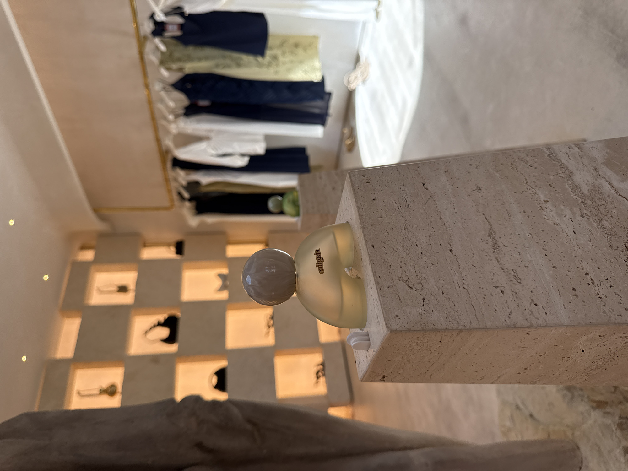 This indoor scene depicts a retail environment, possibly a high-end boutique, though the exact location within Miami, USA, is not discernible from the image alone. A beige, travertine-textured column stands prominently in the foreground, and a frosted glass perfume bottle with a spherical top is mounted on its side. The name "aligala" is inscribed on the bottle.
In the background, a wall adorned with a grid of backlit display niches presents various fashion accessories. A rack of clothing with a range of garments, including white, navy, and neutral tones, is visible in the upper part of the frame. No people are present in the image, and there are no explicit interactions or actions captured. The overall environment suggests a curated and upscale shopping experience.