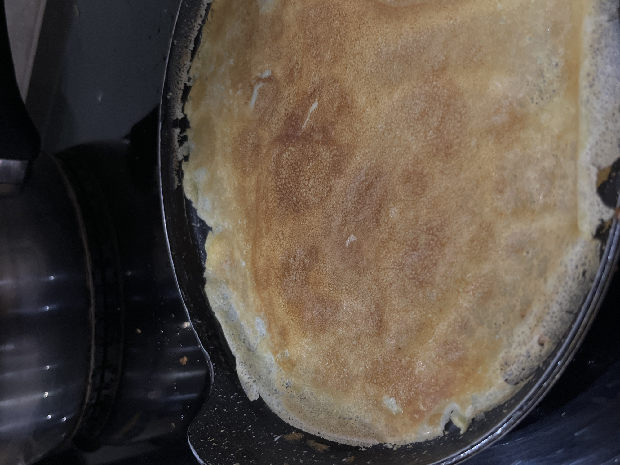 A close-up, overhead shot shows a large, golden-brown crepe cooking in a black frying pan on a stovetop. The crepe is circular and covers most of the pan, with some slightly darker browned areas and lighter, translucent edges. The pan has a speckled pattern on its interior and a black handle. In the background, out of focus, is the shiny surface of a dark pot or pan and a black stovetop. The scene appears to be a domestic kitchen setting. There is no indication of the city of Koroviia, Ukraine in the image.