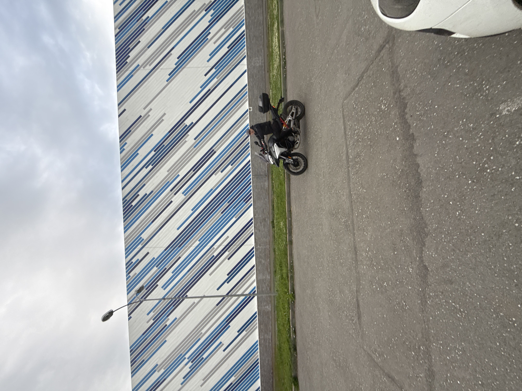 The image shows a person on a motorcycle parked next to a building with a modern, striped facade. The building's design features diagonal lines in varying shades of blue and gray against a white background. To the right of the building is a paved area, likely a road or parking lot, and a strip of green grass.
The motorcycle is dark-colored and appears to be a street bike with a visible storage box mounted on the back. The person is wearing dark clothing and a helmet. They are positioned on the motorcycle, suggesting they are either about to depart or have just arrived.
A street lamp is visible in the lower left portion of the image, extending from the building towards the left. The sky appears overcast, indicating a cloudy day, and the lighting suggests it is daytime.
The overall scene depicts an urban or semi-urban environment, possibly a commercial or industrial area, given the scale of the building and the presence of a road. There is no visible text in the image. The location context provided is Votkinsk, Russia.