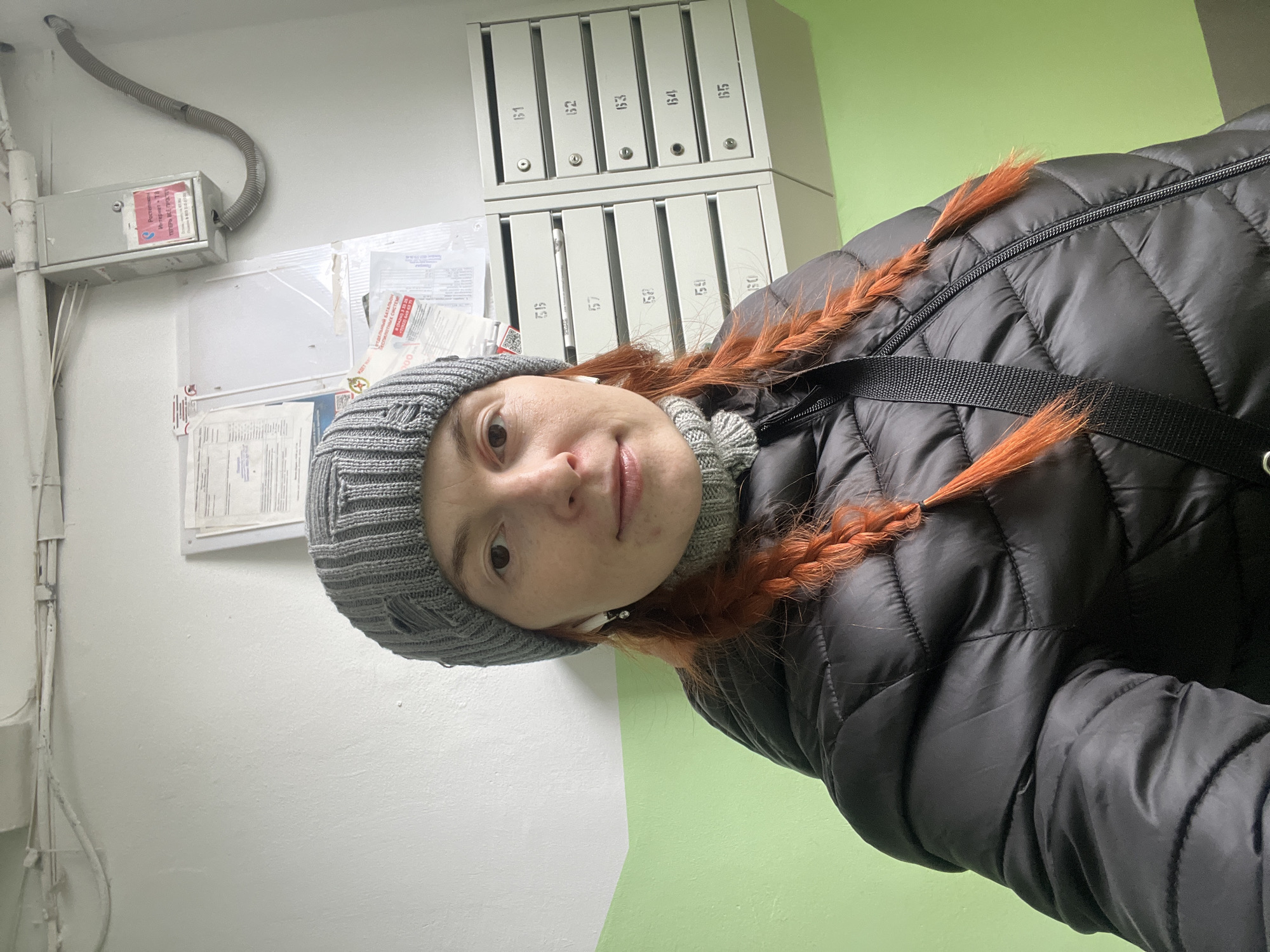 A woman is standing in what appears to be the entrance of an apartment building. She is wearing a gray knitted hat, a gray turtleneck sweater, and a black quilted jacket. Her hair is reddish-brown and braided into two pigtails. A black strap is slung across her body. Behind her is a wall painted white and green, with a row of mailboxes labeled with numbers 56 through 65. Above the mailboxes, there is another row labeled 61 through 65. To the left, there is an electrical box and a notice board with papers and notices affixed to it. The overall setting suggests an interior communal area in a residential building. It is unknown if the location is in the city of Chaikovsky, Russia.