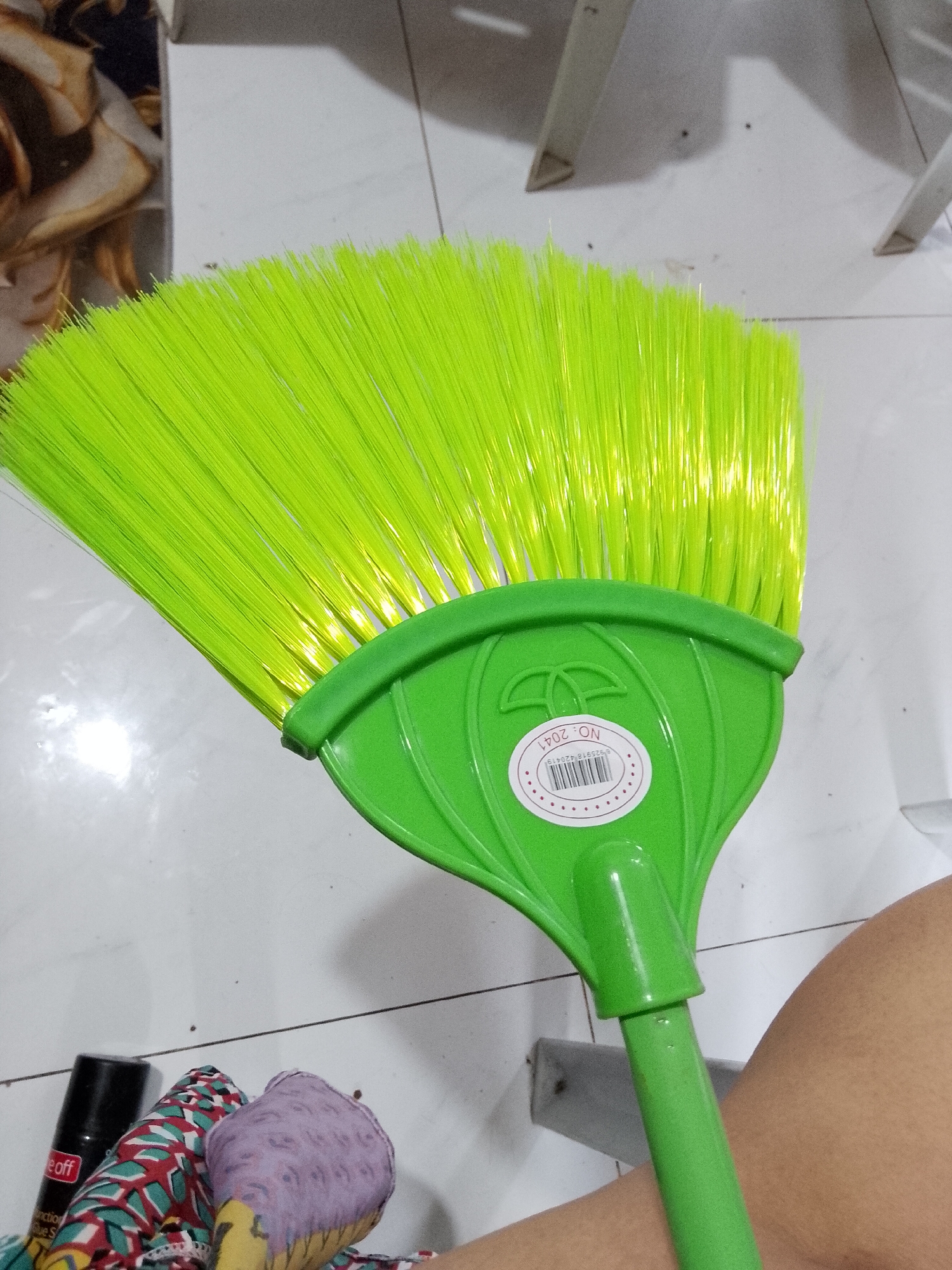 The image captures a close-up view of a bright green plastic broom, held or placed in an indoor setting. The broom features vibrant lime green bristles fanned out, secured by a solid green plastic head. A white, circular sticker is prominently displayed on the broom head, featuring a barcode and the text "NO. 2041" printed upside down. The barcode number "89925918420419" is also visible.
The broom handle, also green, extends downwards, with a portion of it visible in the lower right. On the far right, a section of a person's bare leg or thigh is visible, suggesting the broom is being held or is resting against the person.
The background consists of a light-colored, possibly tiled floor with visible grout lines and some dark speckles. In the lower left, on the floor, are several objects: a black spray can with partially visible text like "eoff", "uction", and "blue S", and some folded textiles or fabric items with colorful patterns, including purple and red-green designs. In the upper background, blurred elements suggest household items, including what appears to be the metal legs of a chair or table and some textured, brownish fabric. The overall scene suggests a domestic environment in Davao City, Philippines. No specific activity is depicted beyond the presentation of the broom. The lighting appears to be ambient indoor light.