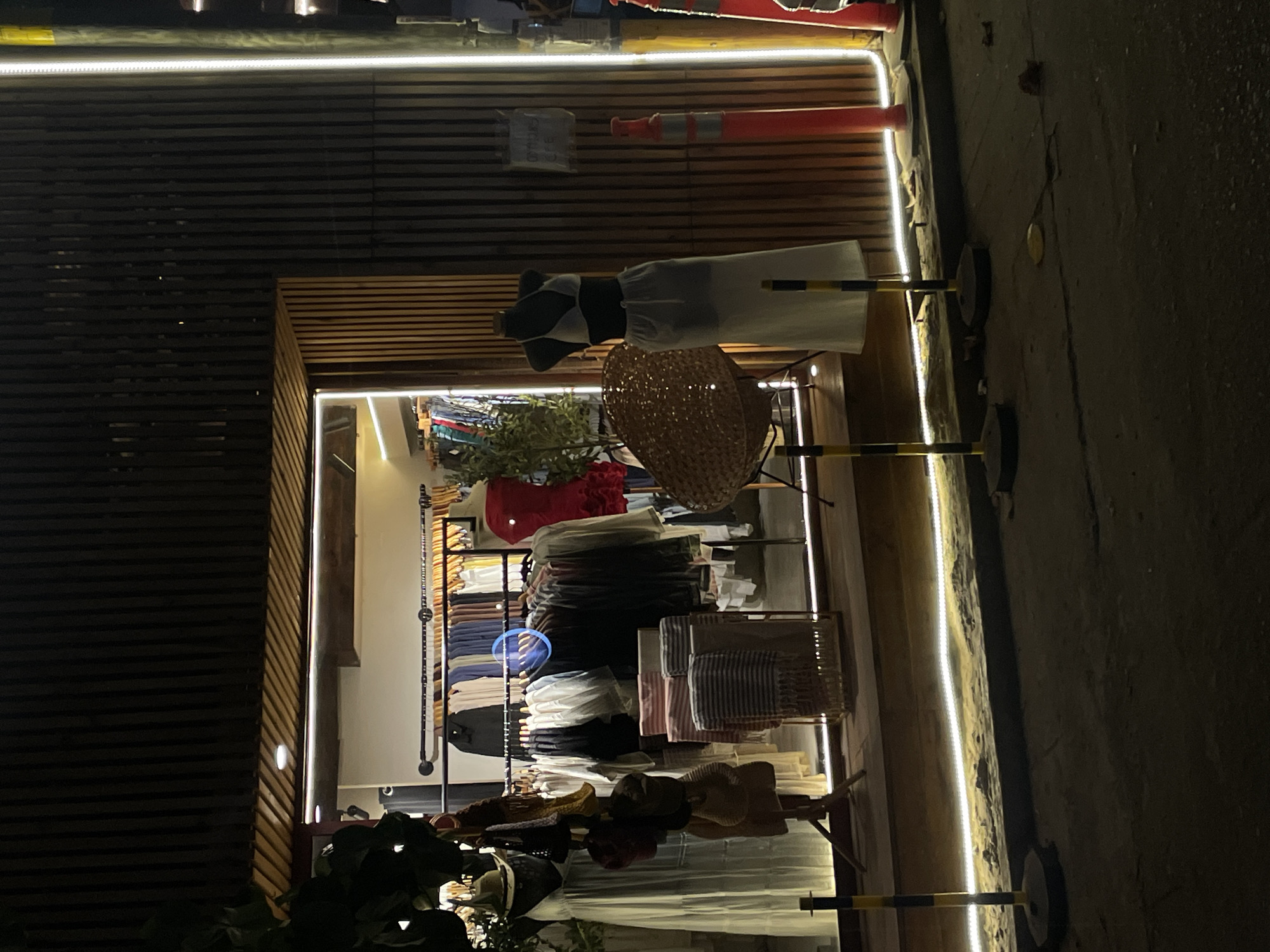 A shop in Brisas de Zicatela, Mexico, displays clothing and accessories. A mannequin dressed in a white dress and black and white top is visible, along with a straw hat. Racks of clothing and hats are on display within the shop's interior. The shopfront is lit with LED strip lights. The exterior is characterized by horizontal wooden slats. Part of the street is visible in the foreground.