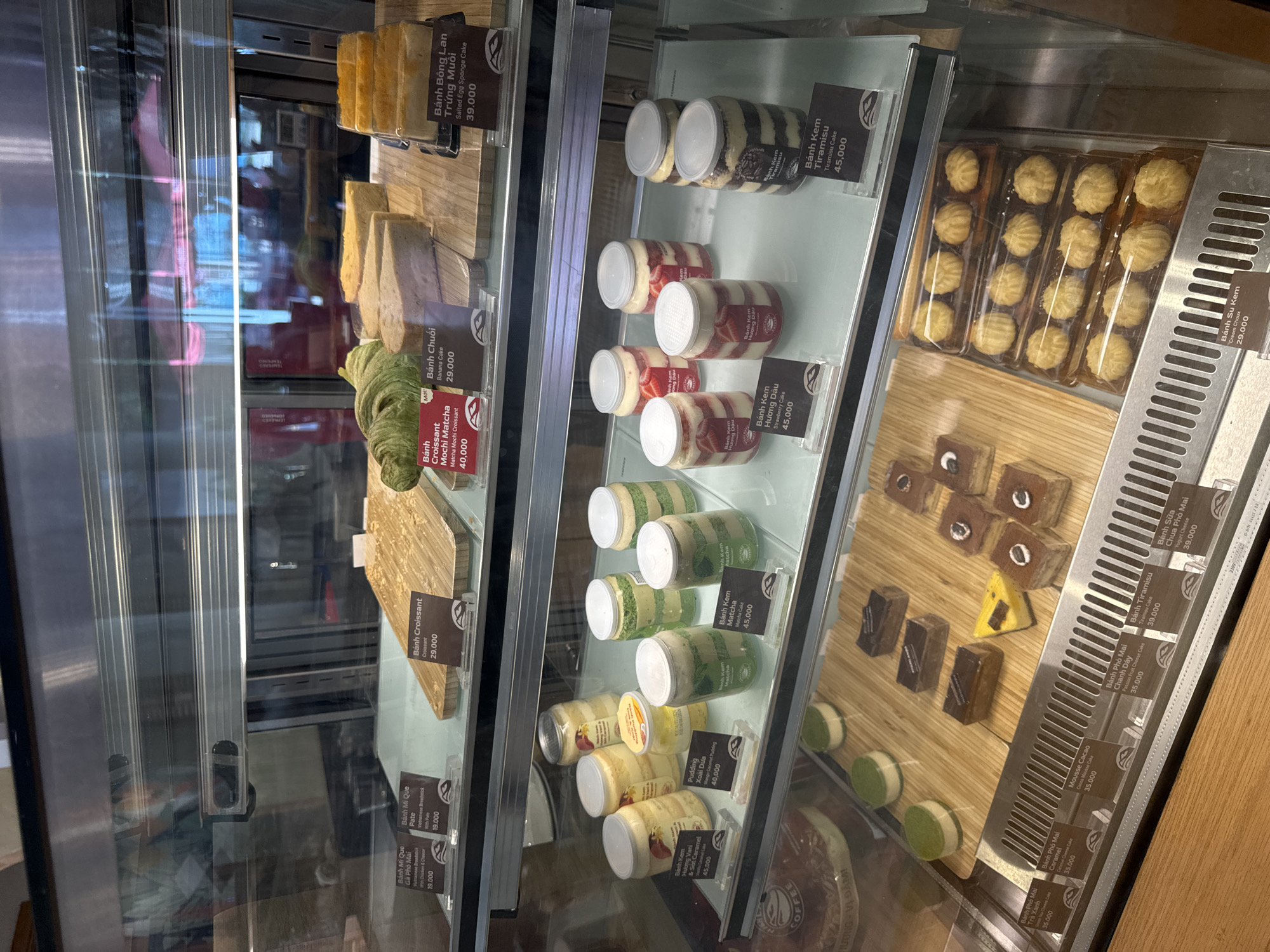 This is a close-up shot of a refrigerated display case filled with various pastries and cakes in a bakery, likely in Phan Thiết, Vietnam. The display case is well-lit, highlighting the tempting treats within. We see a variety of items including sliced cakes, individual dessert cups, and possibly some small pastries.
Notable items include several layers of sliced cakes that appear to be sponge cake varieties, some with visible fruit fillings. There are also numerous small, individual dessert cups, some layered with cream and what looks like fruit, others with a green hue suggesting matcha flavor. The labels indicate a range of products with Vietnamese names and prices in Vietnamese Dong. Some of the visible text includes "Bánh Bông Lan Trứng Muôi" (Salted Egg Sponge Cake), "Bánh Kem Matcha" (Matcha Cake), "Pudding Xoài Dưa" (Mango Coconut Pudding), and "Bánh Tiramisu" (Tiramisu Cake). There are also labels for "Bánh Croissant" (Croissant) and "Bánh Mì Que" (Breadstick).
On a wooden platform to the right, a selection of small, rectangular chocolate cakes with a white topping are neatly arranged, alongside some round, green-colored desserts. Further to the right, there are trays of small, round cookies or biscuits. The overall impression is that of a well-stocked and appealing patisserie. The reflections in the glass show the interior of what appears to be a shop, but no people are clearly visible. The lighting suggests it's daytime, likely during business hours.