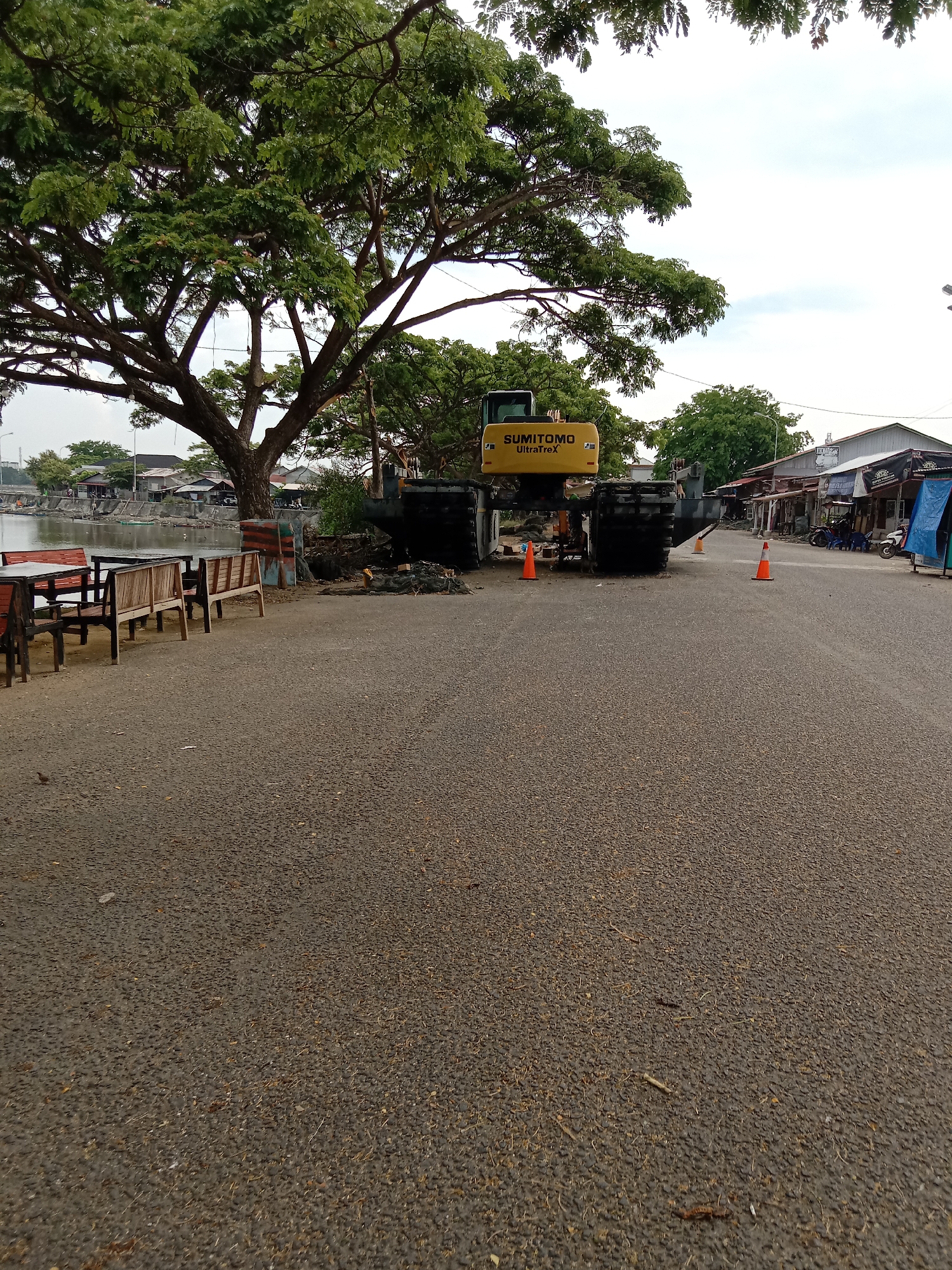 The image depicts a street scene in Lhokseumawe, Indonesia. A large yellow Sumitomo UltraTrex excavator is positioned on the asphalt road, partially obstructing traffic. Two orange traffic cones are placed in front of the excavator, likely for safety. To the left of the excavator, there is a tree and several wooden benches and tables. Beyond the benches, there is a body of water, possibly a river or lake, with buildings visible in the background. On the right side of the road, there are buildings with shops, scooters, and people, suggesting a commercial area. The sky is overcast. The road surface is worn and has some scattered debris.