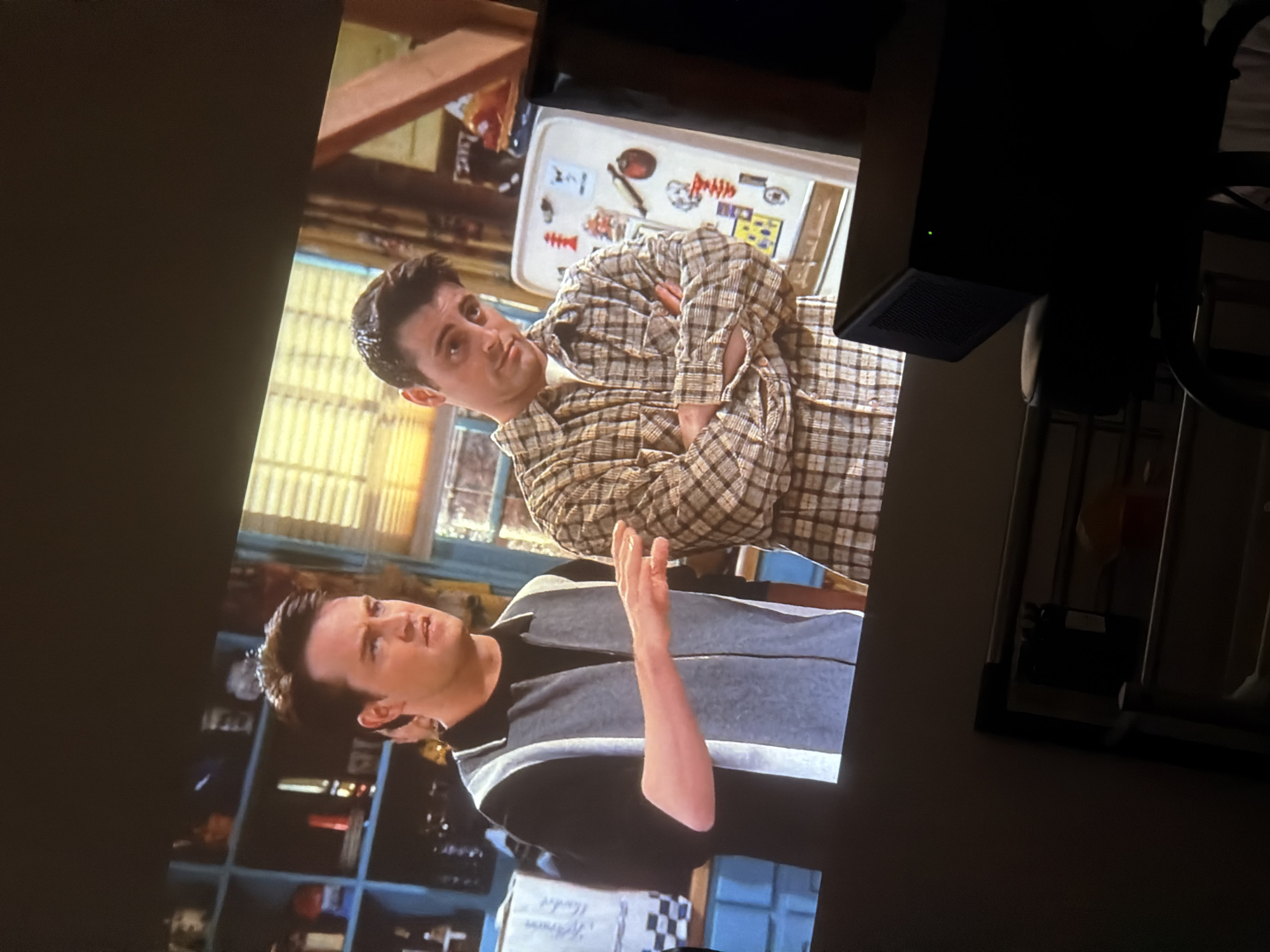 The image shows a TV screen playing an episode of the sitcom "Friends." In the episode, two male characters are visible: one wearing a black and gray bowling-style shirt, gesturing with his right hand, and the other wearing a brown plaid pajama set, with his arms crossed. The scene appears to be taking place in Monica and Rachel's apartment. A refrigerator is visible in the background, covered with magnets. To the right of the TV screen, there is a black electronic device sitting on a metal shelf. The location of the image cannot be determined, and the city of Cape Town, South Africa is not related to the image.