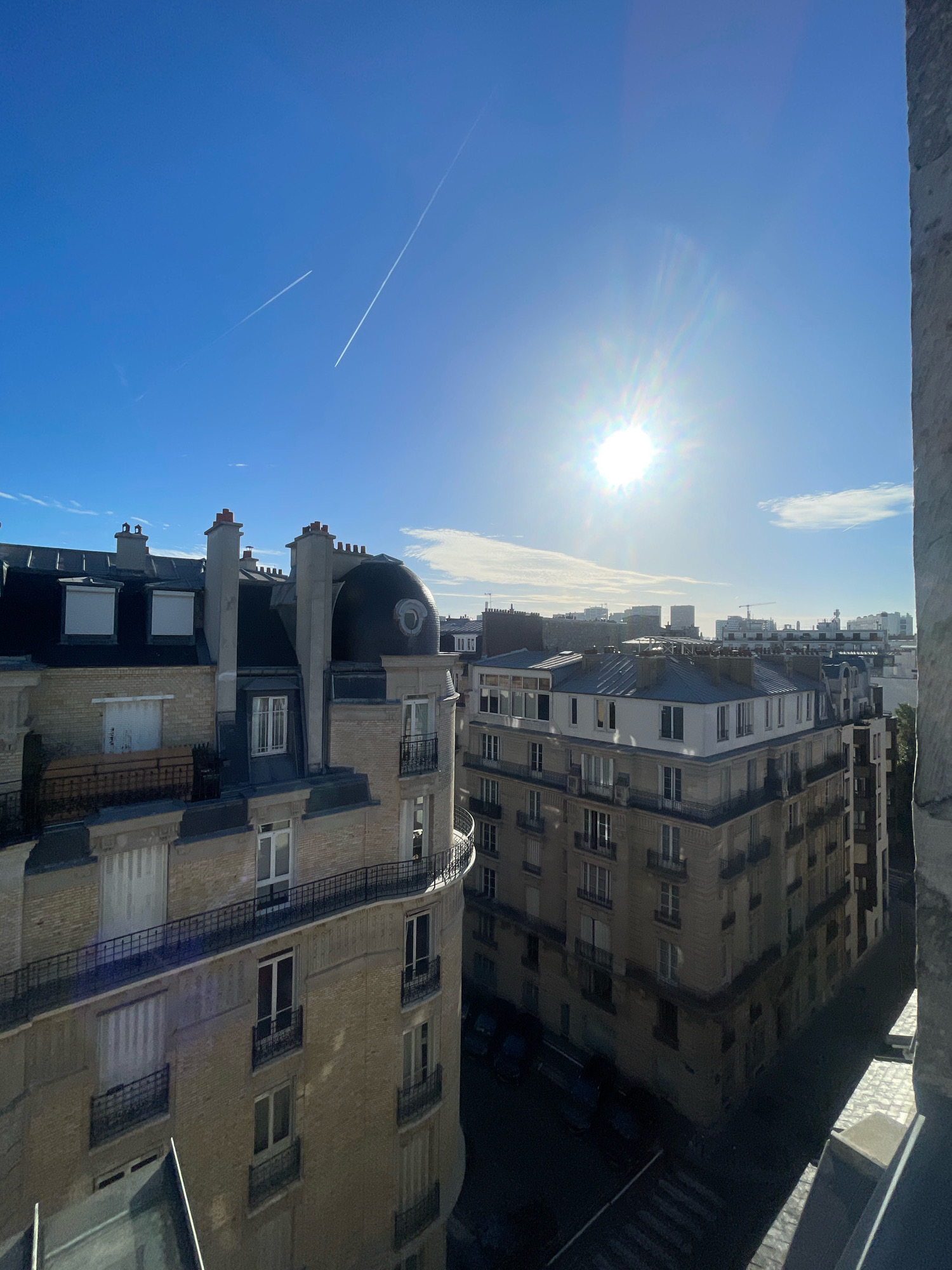 The photograph presents a high-angle view of Parisian rooftops under a clear blue sky with a bright sun. Two distinct contrails from airplanes streak across the upper portion of the sky. The foreground is dominated by the rooftops and facades of traditional Parisian apartment buildings. One building, with a light-colored brick facade, features several windows, balconies with black railings, and chimneys. A distinctive domed architectural feature is visible on this building's corner. Adjacent to it, another building with a darker, tiled roof and multiple windows is visible. The mid-ground shows a dense urban landscape with more rooftops and buildings, some of which appear to be under construction, indicated by cranes in the distance. The sun is high and bright, casting strong shadows and creating lens flare. The street level is mostly in shadow, with parked cars visible in some areas. The image captures a sunny day in Paris, likely in a residential area.
