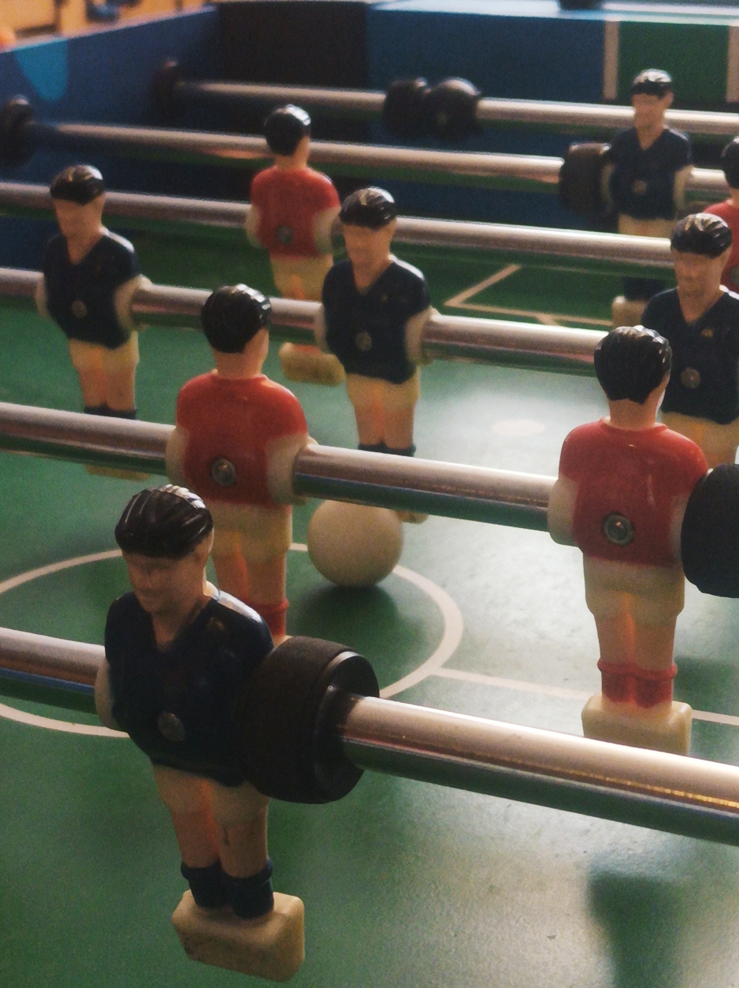 The image shows a close-up of a foosball table. The table is green with white lines marking the playing field. Two teams of foosball players are visible, one in navy blue and the other in red. The players are attached to metal rods. A white foosball sits on the field between two rods. The image does not provide any information about the location of the foosball table, therefore, it is not possible to establish if it is in Porlamar, Venezuela.