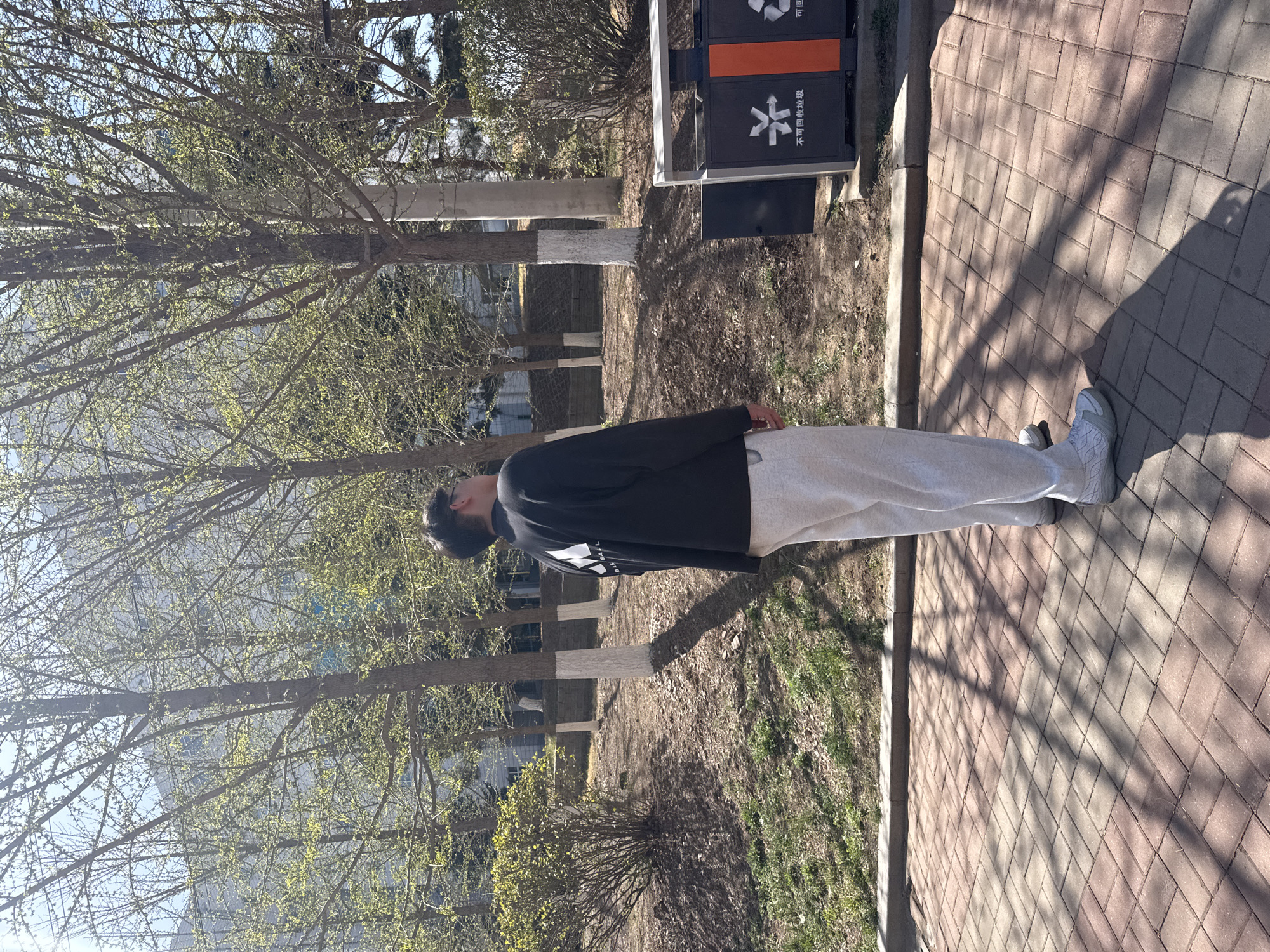 The image shows a person in Zhongtun, China standing on a brick walkway near a curb. They are wearing a dark long-sleeved shirt and light-colored sweatpants and shoes. Behind them are trees with budding leaves and a recycling/trash bin. The ground around the bin is covered with dirt and sparse vegetation. Some tree trunks are painted white at the base. In the background, there are buildings partially obscured by the trees. The overall environment suggests a park or green space within an urban setting.