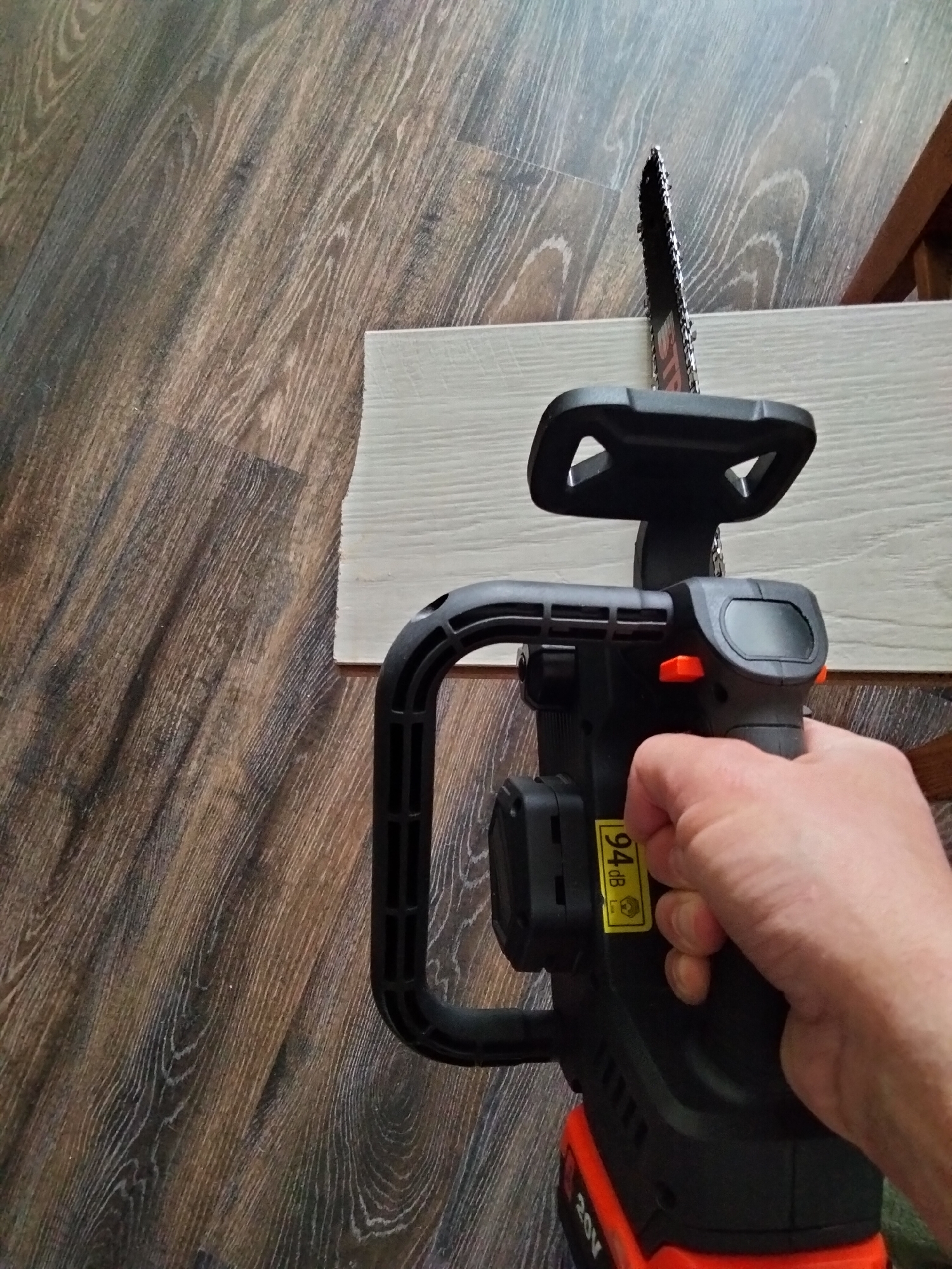 An overhead shot of a person holding a black and orange cordless chainsaw with an orange handle and battery pack, placed on a wooden floor. The chainsaw is resting on a light-colored plank of wood. A yellow sticker with "94 dB" is visible on the chainsaw. The background shows a wooden floor with a distressed wood grain pattern.