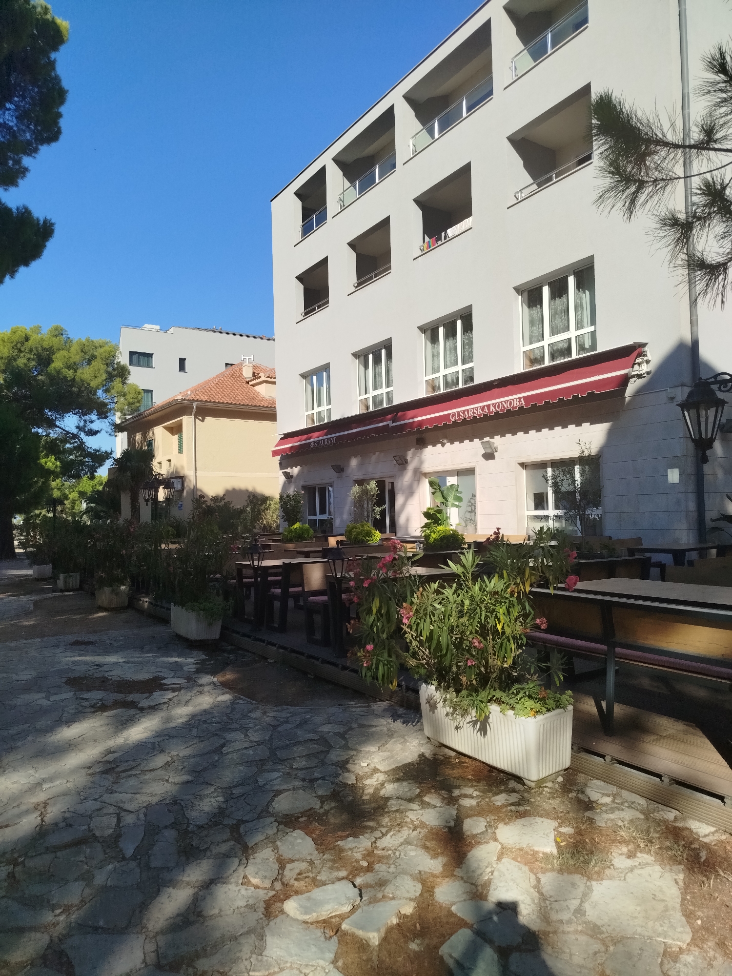 The image captures an outdoor seating area of a restaurant with a red awning that reads "GUSARSKA KONOBA". The restaurant appears to be on the ground floor of a modern, light-colored building with balconies. In front of the restaurant, there are several tables and chairs set up for dining, surrounded by potted plants and greenery. The ground is paved with irregular stone slabs, with shadows cast by trees. To the left of the restaurant, there is another building with a terracotta roof, and further in the background, a taller, grey building. The sky is clear and blue, indicating a sunny day. A pine tree's branches are visible in the upper right corner, partially obscuring the view of the building.