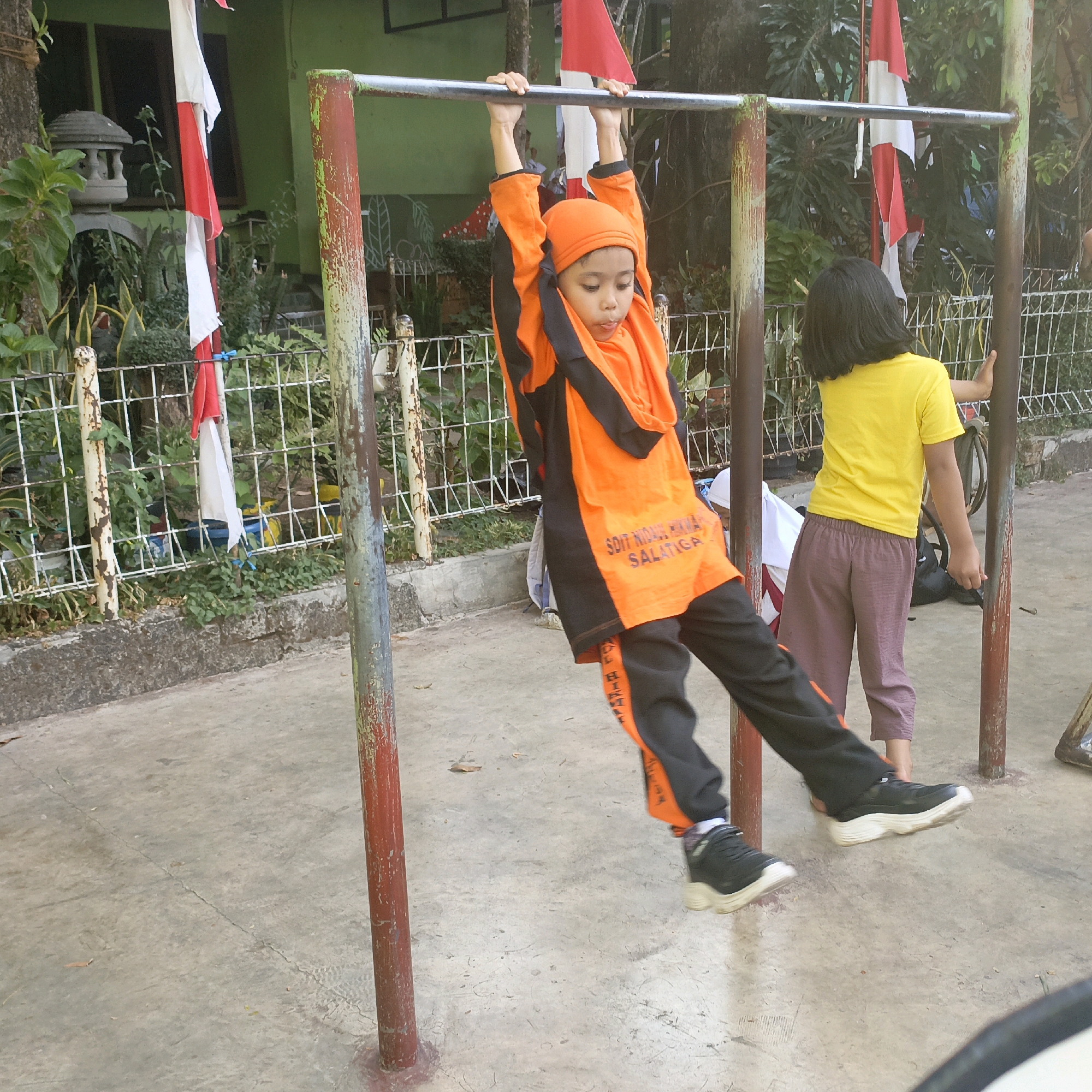 In Salatiga, Indonesia, at ground level, a child dressed in an orange and black sports outfit, including an orange head covering, is suspended from a horizontal bar, likely a pull-up bar. Their body is angled downwards, and their hands grip the bar. Their legs are extended, and their feet are in motion, with black sneakers featuring thick white soles. The background reveals a green building with two dark-windowed sections. A white and red flag, possibly the Indonesian flag, is visible, draped on a pole. Another child, seen from the back, is wearing a yellow t-shirt and brown pants, standing near the bar and touching one of its vertical supports. The ground is a concrete surface with visible cracks. A white metal fence with vertical posts and horizontal bars borders the area. Lush green vegetation is present behind the fence, interspersed with ornamental plants.