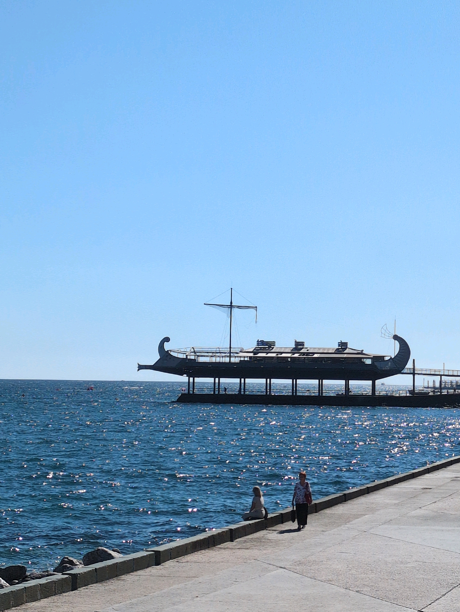 A wide shot shows a bright blue sky over a calm, sparkling blue ocean. In the middle ground, a long, dark structure resembling an ancient galley ship is elevated on stilts over the water. The ship has a curved, pointed prow and stern, and a mast with rigging. It appears to be a pier or building.
In the foreground, a paved walkway runs along the edge of the water. Two people are present on the walkway. One person, dressed in light-colored clothing, is sitting on the edge of the walkway facing the water. Further along the walkway, another person, wearing a patterned top and dark pants, is walking away from the camera. The ocean has small waves that catch the sunlight, creating a shimmering effect. The walkway is bordered by large stones and concrete. The scene appears to be a sunny day in a coastal area.