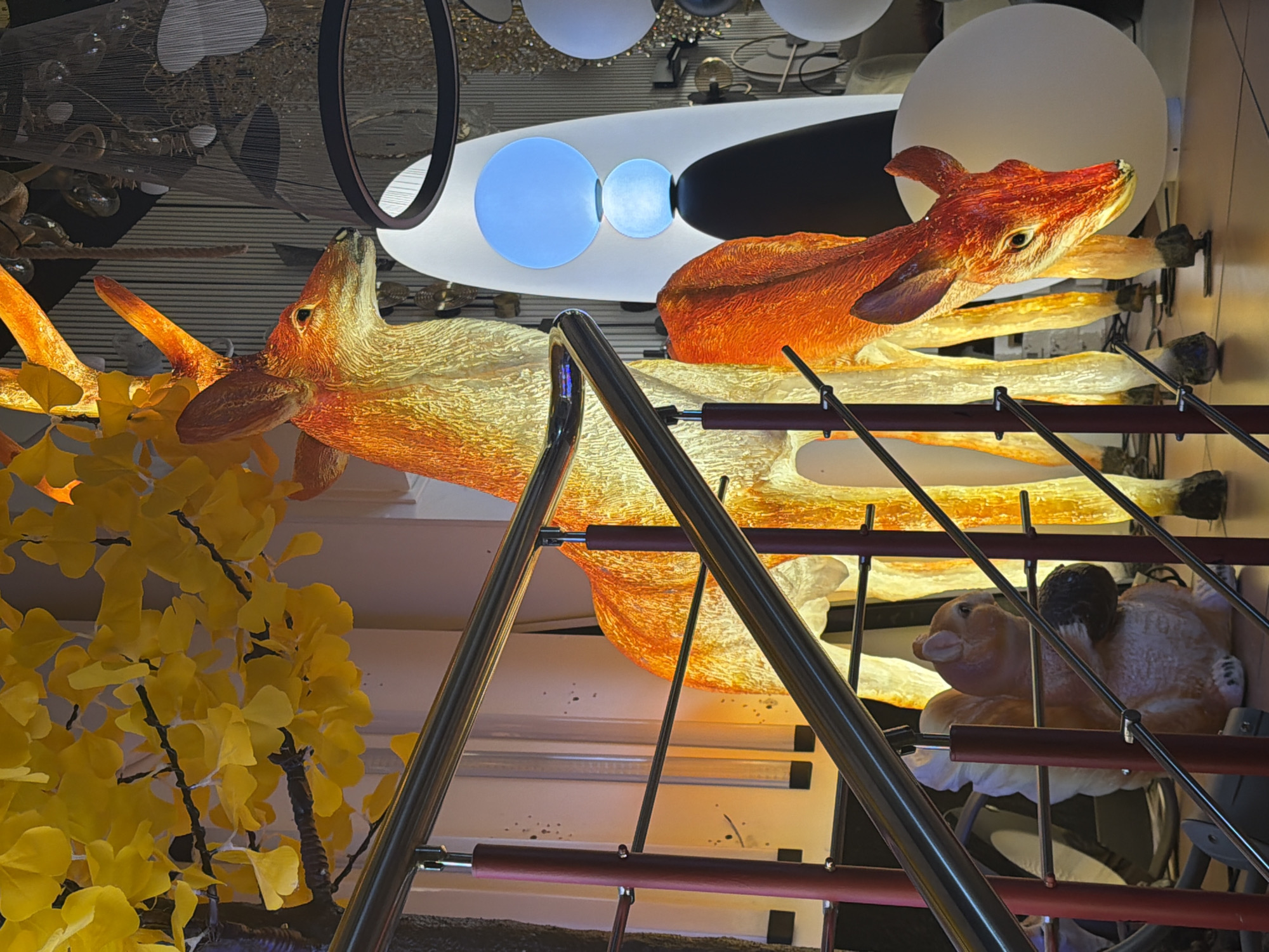 The image depicts a decorative display featuring two illuminated deer sculptures, one larger and one smaller, positioned near a staircase. The deer are rendered in warm orange and yellow tones, suggesting they are lit from within or by external lighting. A squirrel sculpture holding a nut is visible beneath the staircase railing. In the background, various modern light fixtures are displayed, including spherical and ring-shaped designs, some of which are illuminated. A branch with yellow leaves, possibly artificial, hangs from the upper left of the frame. The overall setting appears to be a retail environment, possibly a showroom for lighting or home decor. The specific location in Moscow, Russia, is not discernible from the image itself.