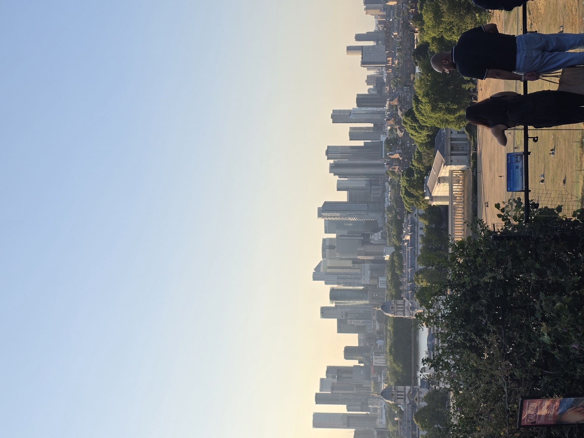 This is a photograph taken from a high vantage point overlooking a cityscape. In the foreground, two individuals, a woman and a man, are standing and looking out at the view. The woman, on the left, has dark hair and is wearing a black top and a backpack. The man, on the right, is bald, wearing a black polo shirt and light-colored trousers, and is holding a large tote bag. A railing separates them from the expansive view.
In the midground, there is a large, open grassy area where several people are scattered, some sitting, others cycling. Beyond this parkland, several notable buildings are visible. Two prominent, domed structures with classical architecture are situated near a body of water. Further in the distance, a dense cluster of modern skyscrapers dominates the horizon, characteristic of a central business district. The sky is clear and light blue, indicating a daytime scene, likely late afternoon or early evening due to the warm, soft light.
The overall scene suggests a panoramic view of London, United Kingdom, possibly from Greenwich Park, offering a perspective that combines historic architecture with the modern skyline of Canary Wharf in the distance.