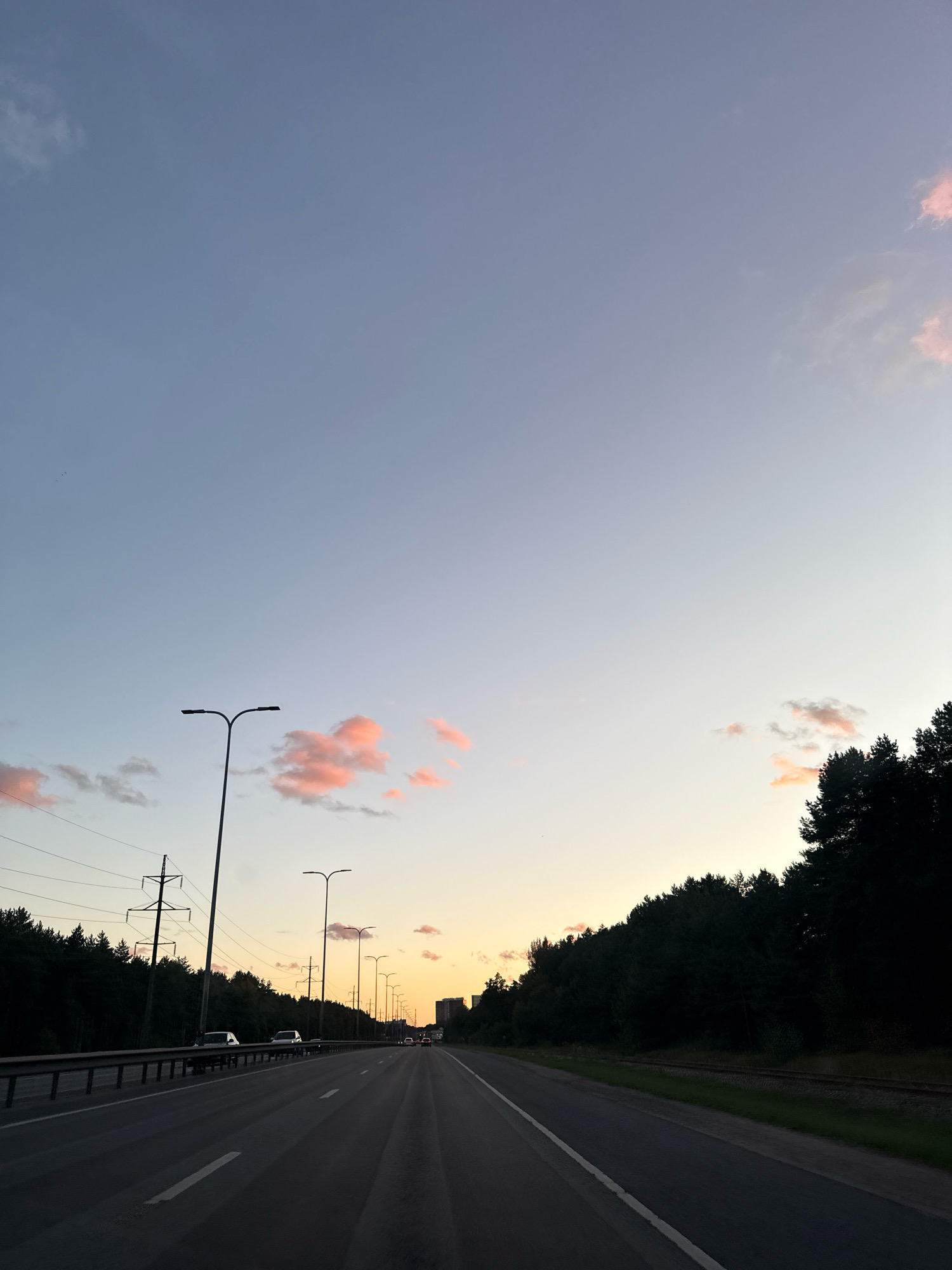 The image depicts a highway in Tallinn, Estonia, at dusk. The sky is a gradient of pale blue at the top, transitioning to a soft orange and yellow hue near the horizon, with scattered pink clouds. The highway stretches into the distance, with multiple lanes marked by white lines. Several cars are visible on the road. On the left side of the highway, there is a metal guardrail and a line of tall streetlights. Beyond the guardrail, a dense forest is visible, interspersed with power lines and utility poles. On the right side of the highway, another dense line of trees forms a dark silhouette against the twilight sky. In the far distance, some buildings can be faintly seen. The overall atmosphere is serene and captures the transitional light of early evening.