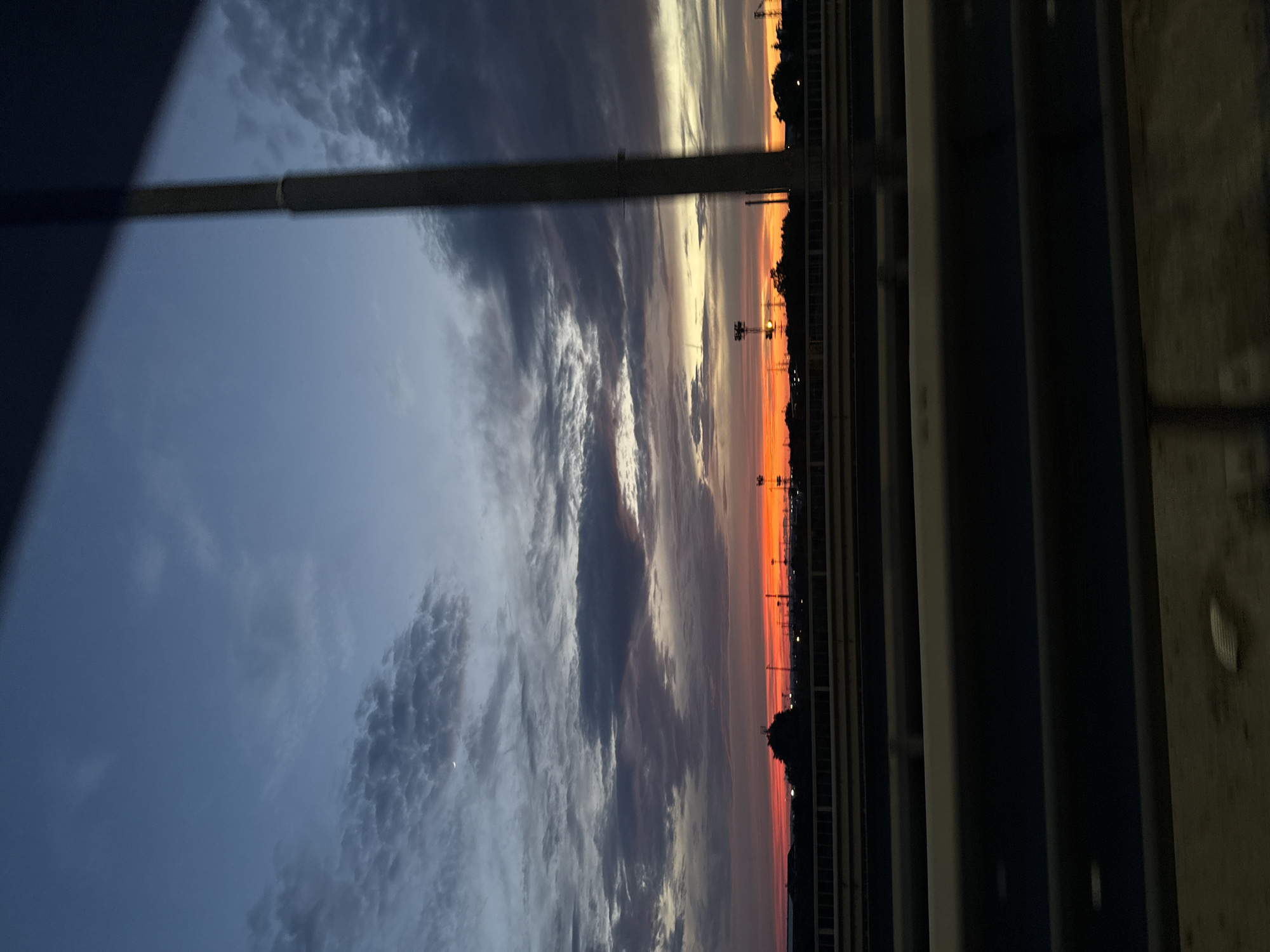 The image depicts a view of the sky at dusk, likely from a moving vehicle on a highway in Burgas, Bulgaria. The sky exhibits a gradient of colors, transitioning from a deep blue at the top to vibrant oranges and reds near the horizon. Wispy clouds are scattered across the sky, some tinged with the sunset colors. A sliver of the moon is visible in the upper left quadrant. In the distance, silhouetted against the bright horizon, are structures that appear to be industrial cranes and possibly light poles. A dark silhouette of trees and buildings is visible along the horizon line. In the foreground, there are blurred horizontal elements, likely guardrails or barriers of the highway, suggesting the perspective of someone looking out from a vehicle. The overall scene captures a moment of transition from day to night, characterized by dramatic sky colors and the distant industrial landscape.