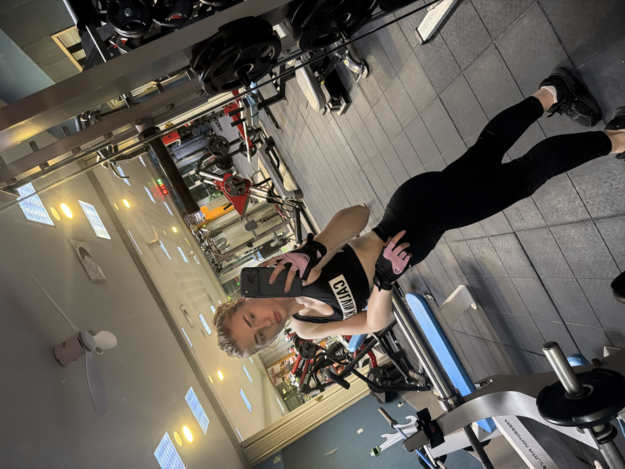 A young woman with blonde hair tied up in a bun takes a mirror selfie in a gym. She is wearing a black sports bra with "CALVIN" written on it in white text, black leggings, and black sneakers with white socks. She is also wearing pink fingerless gloves. The gym appears to be well-equipped with various weight machines and equipment. The lighting is bright, with fluorescent lights on the ceiling and a clock displaying "01:51" visible in the background. The floor is tiled with black rubber mats.