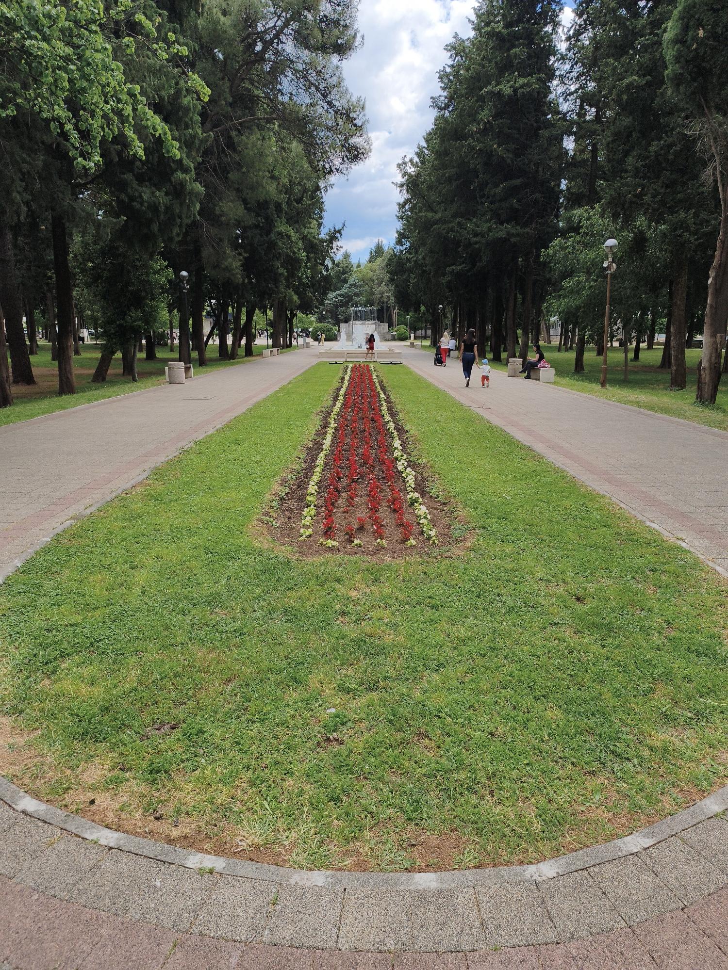 The image depicts a park in Trebinje, Bosnia and Herzegovina, featuring a long, rectangular grassy area in the foreground that narrows towards the background. A bed of red and white flowers lines the center of the grassy area. A pedestrian walkway runs along either side of the grass, with trees lining both sides of the walkways. People are walking along the walkway in the background, including a woman and a child. Benches are placed along the walkway, and some people are sitting on them. In the far background, a fountain is visible. The weather appears to be partly cloudy.