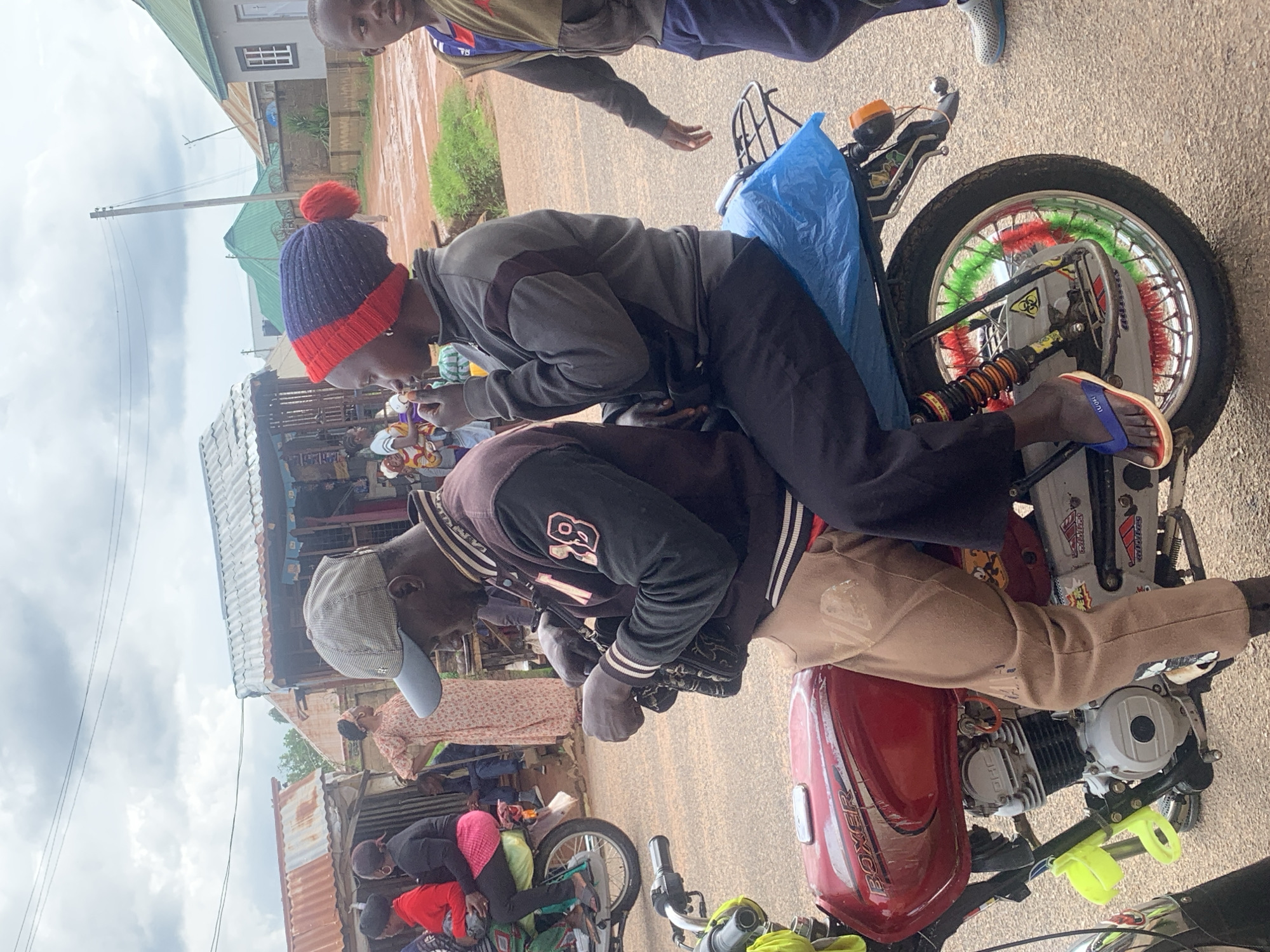 The image shows two young men on a motorcycle parked outside a street-side stall in Barakin Akawo, Nigeria. The man in the front is wearing a blue and red knitted beanie and a grey jacket. He is looking down and appears to be eating or holding something. The man behind him is wearing a black jacket with white stripes and a beige beanie. He is also looking down. The motorcycle is red and has colorful decorations on its spokes. Behind the motorcycle, there is a stall with corrugated metal roofing and various items for sale. Other people are visible in the background, including women and children. The ground is dusty, and the sky is overcast, suggesting it might be a cloudy day. The overall scene depicts a common everyday moment in a Nigerian street market. Some visible text includes "BOXER" on the motorcycle's fuel tank and other decals.