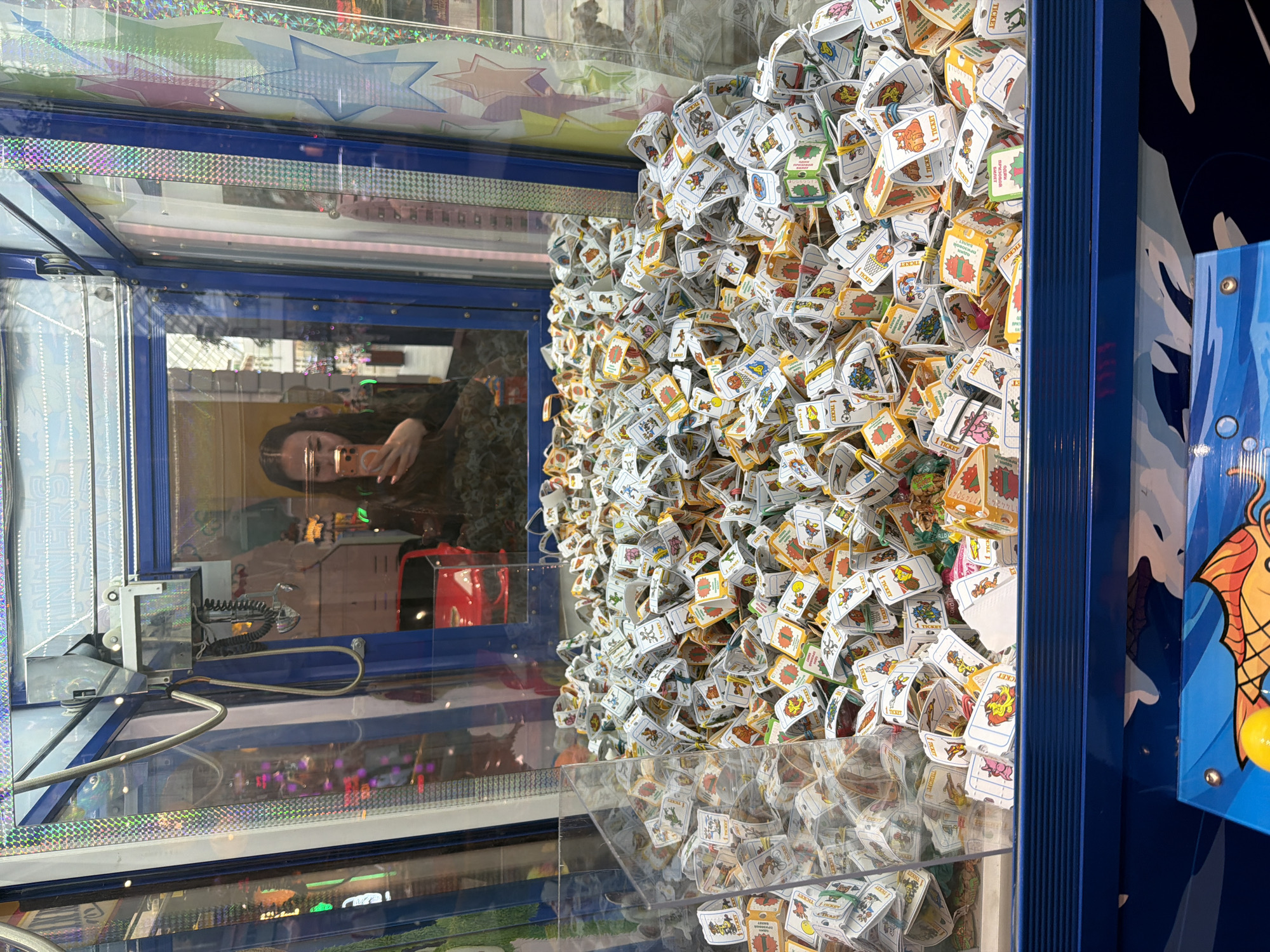 The image shows the inside of a claw machine, filled with small, cube-shaped prizes. Each prize is wrapped in white paper with colorful cartoon illustrations and text, some of which appear to be "1 TICKET." The prizes are densely packed, forming a large mound within the transparent enclosure of the machine. In the background, the reflection in the glass shows a woman holding a smartphone, presumably taking a selfie. Other reflections include lights and what appears to be a red car. The machine itself has blue accents and decorative elements along the top and bottom. The overall scene suggests an indoor amusement or entertainment setting.