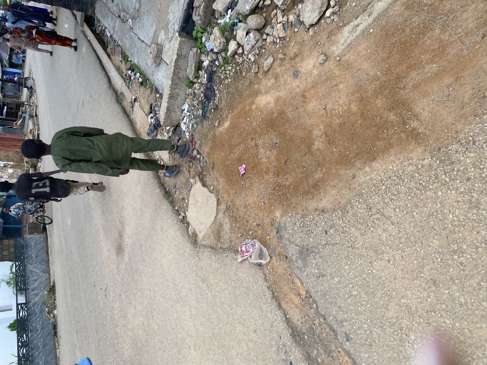 In Jos, Nigeria, a young boy in a green kaftan walks away from the camera on a paved road next to a dirt drainage ditch filled with debris and rocks. To his left, another boy wearing a black t-shirt with the number 13 walks away from the camera. Further in the background, several people are walking on the road, and a child is riding a bicycle. The environment appears to be a street scene with buildings on either side. There is trash and debris scattered along the side of the road and in the drainage ditch.