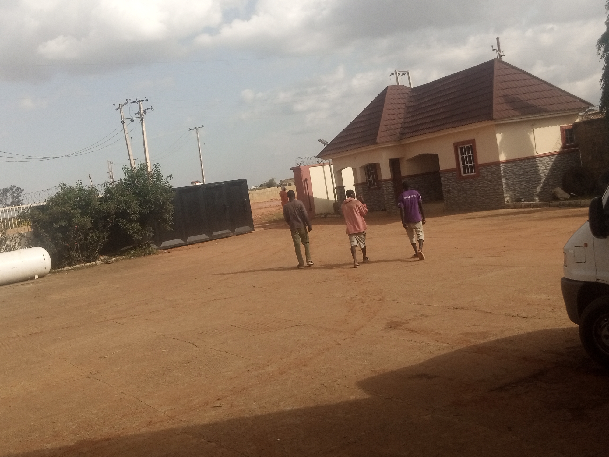 A group of three men are walking away from the camera across a dusty, reddish-brown surface. The man on the far left wears a gray long-sleeved shirt and green pants. The middle man is dressed in a pink long-sleeved shirt and light-colored shorts. The man on the right wears a purple long-sleeved shirt with white text and light-colored pants.
Behind the men, a building with a brown tiled roof and cream-colored walls is visible. The lower part of the building's exterior is adorned with gray stone-like tiles. A dark metal gate stands to the left of the building. In the background, power lines and poles suggest an outdoor, possibly rural or semi-urban setting. A white cylindrical object, possibly a tank, lies on the ground to the far left. A white van is partially visible on the right side of the image.
The sky is partly cloudy, indicating it is daytime. The overall lighting suggests it could be late morning or early afternoon. There is no clear indication of any specific event or strong emotions. The text on the purple shirt is not legible.