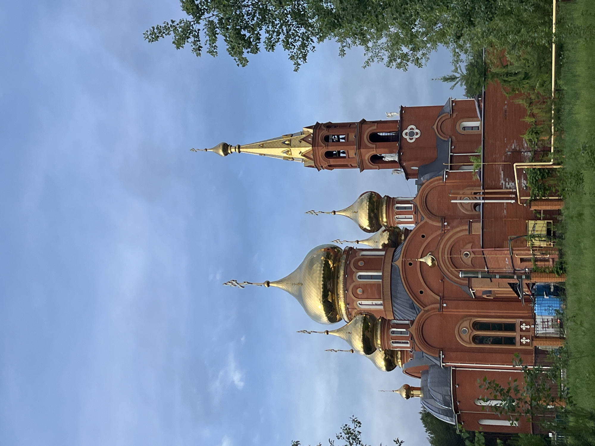 The image depicts the domes and structure of the Church of the Holy New Martyrs and Confessors of Russia, located in the city of Chaikovsky, Perm Krai, Russia. The church features multiple golden domes with crosses, and a prominent spire. The building's exterior is constructed of reddish-brown bricks with architectural details such as arched windows and decorative elements. Green foliage is visible along the edges of the frame, suggesting the church is surrounded by trees or landscaping. The sky is blue with some clouds.