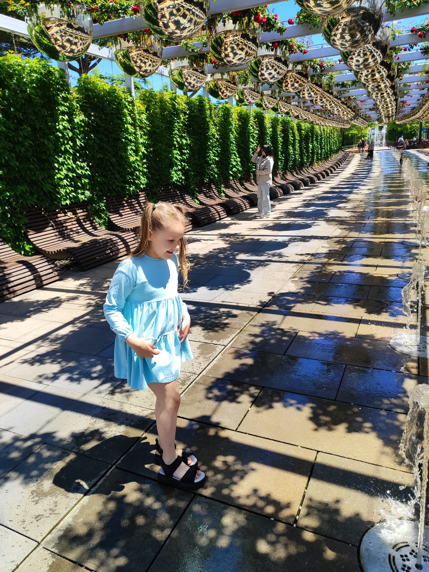 This media file captures a vibrant outdoor scene within a modern park or promenade in Krasnodar, Russia, on a bright, sunny day. The dominant feature is a long walkway, partially covered by a white pergola-like structure. This structure is adorned with numerous large, reflective, spherical ornaments that cast distorted reflections of the surroundings, including the blue sky, green foliage, and red flowers growing along the pergola.
To the left of the walkway, a tall, continuous wall of lush green ivy provides a verdant backdrop, fronted by a series of long, slatted wooden benches that curve gently. Dappled shadows from the overhead structure and foliage are cast across the light-colored paved walkway, indicating strong sunlight.
In the foreground, a young girl with light brown hair styled in pigtails, wearing a light blue, long-sleeved dress and black sandals, is the primary subject. She is standing on the paved path, looking downwards and slightly to her right, seemingly engrossed by one of the small geyser-like fountains that line the right side of the walkway. These fountains spray water upwards, creating wet patches on the pavement. The girl appears to be curiously observing or interacting with the water.
Further down the walkway, an adult female in light-colored clothing is visible, standing and looking towards the right. Other indistinct figures can be seen walking in the distance, adding to the leisurely atmosphere. The overall impression is one of a well-maintained, artistic, and engaging public space. Faint, unreadable text is visible on the circular grates of the floor fountains.