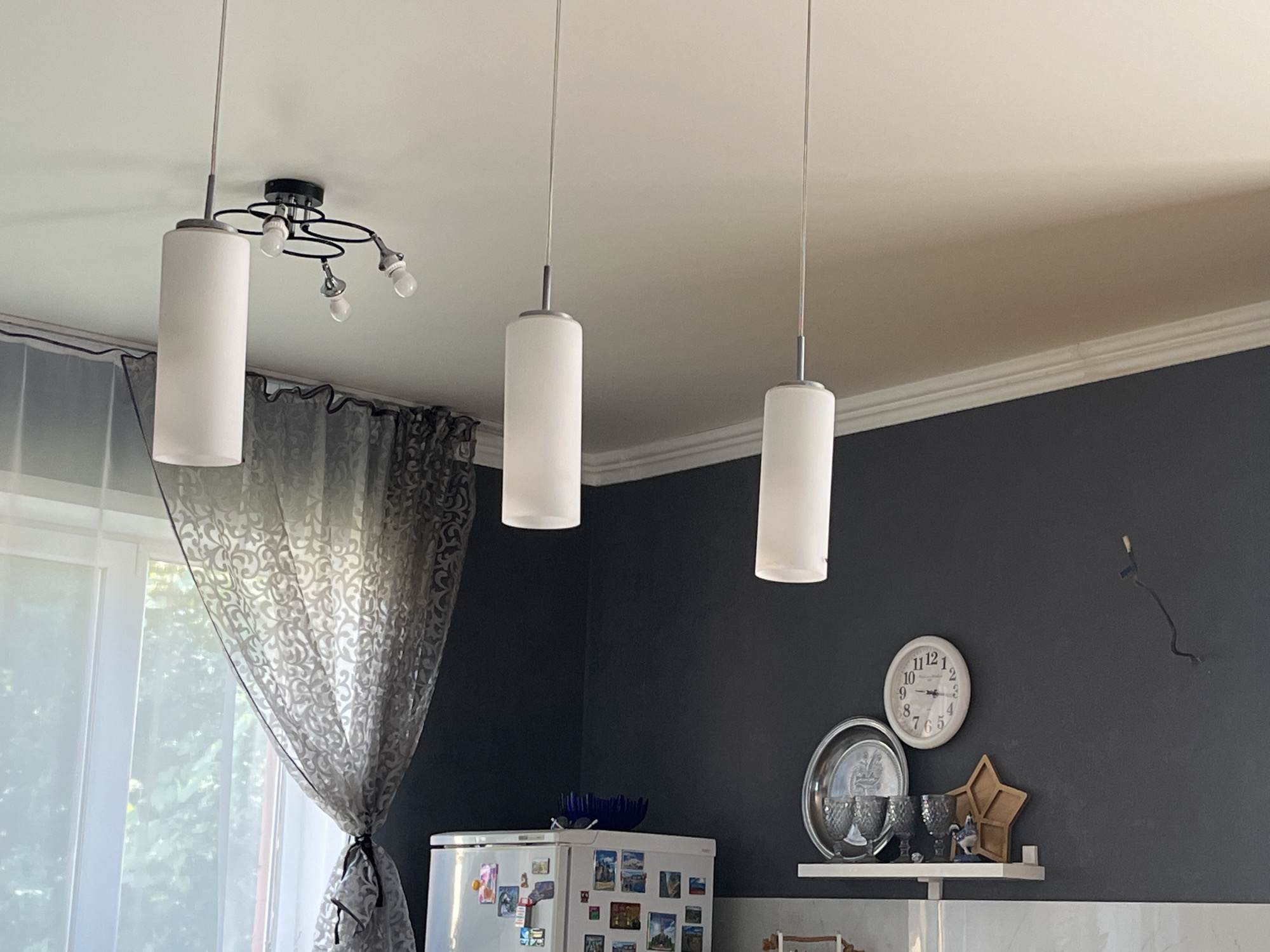 The image depicts an interior scene, possibly a kitchen or dining area, with dark gray walls. Three cylindrical pendant lights hang from the ceiling. To the left, sheer curtains with a swirling pattern are drawn across a window. A refrigerator adorned with magnets is visible in the lower left. On the wall, a clock shows the time as approximately 10:08. Below the clock, a white shelf holds several items, including decorative glasses, a small figurine, and a star-shaped object. A loose black wire hangs from the wall above the shelf. There is no indication of the city of Saratov, Russia, within the image.