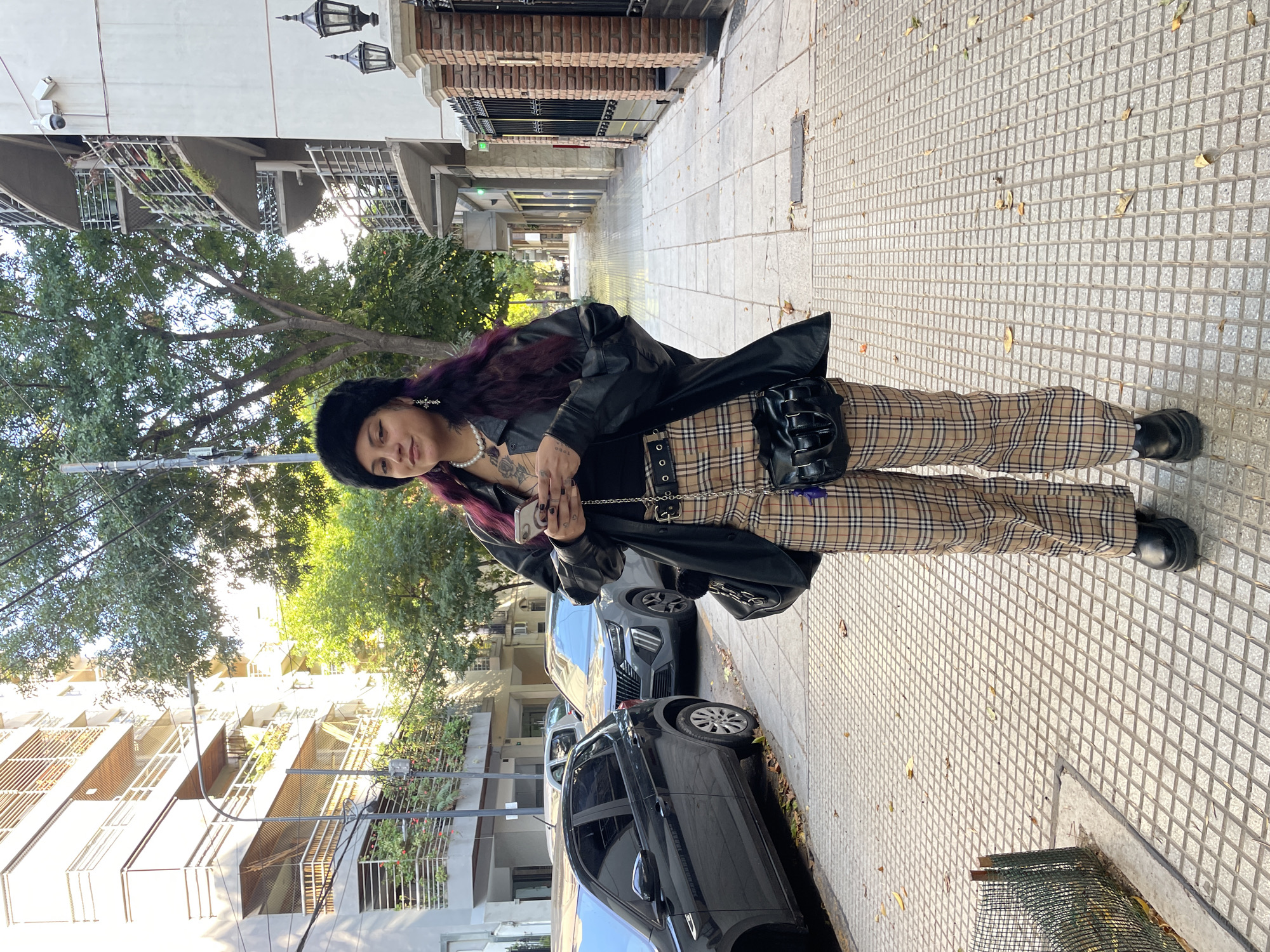 A young woman stands on a sidewalk in a residential area of Buenos Aires, Argentina. She wears a black furry hat, a black leather jacket, a pearl necklace, and tan plaid trousers with a studded belt. She also has visible tattoos. She holds a smartphone in her hands, looking down at it. Behind her are parked cars and apartment buildings with trees lining the street. The sidewalk is paved with square tiles. Overhead streetlights are visible in the distance.