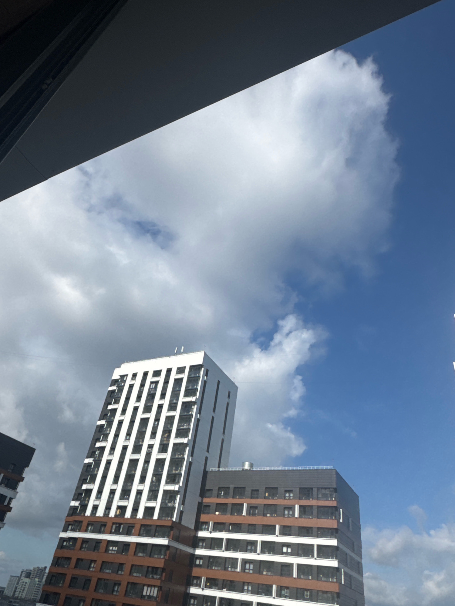 The image depicts a view of modern apartment buildings in Novosibirsk, Russia, against a partly cloudy blue sky. In the foreground, a dark overhang, possibly a balcony or part of another building's structure, occupies the upper left portion of the frame, casting a shadow. Below this, two prominent residential buildings are visible. The taller building, on the left, features a striking facade with a repeating pattern of white vertical elements and dark windows. Its base is clad in a reddish-brown material, interspersed with dark windows. To its right, a second building, which appears to be connected or adjacent, has a similar architectural style with horizontal bands of dark brown and white, and numerous windows. The sky is a vibrant blue with large, fluffy white clouds, and some areas of darker, more diffused clouds, suggesting dynamic weather. The overall scene conveys an urban residential landscape under a bright sky.