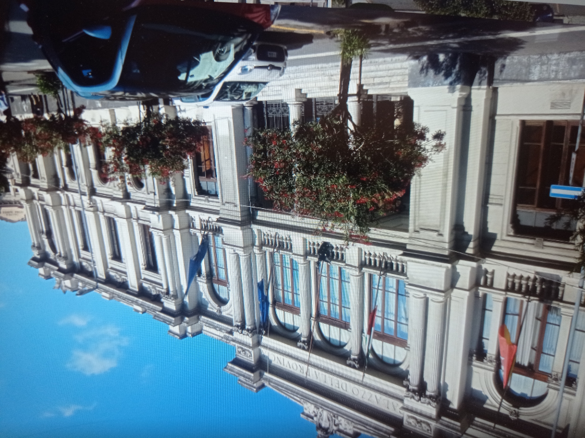 The image shows a large, ornate building with classical architecture. The building appears to be made of light-colored stone and features arched windows, columns, and a balustrade. Flags are visible on poles outside the building. In front of the building, there are trees with red flowers, and several cars are parked on the street. The sky is blue with a few scattered clouds.
The text above the windows on the building reads "PALAZZO DELLA PROVINCIA". This text suggests that the building is a provincial administrative building, likely in Italy. There is no indication that this image is related to the city of Mukachevo, Ukraine. The architectural style, the language on the building, and the presence of Italian flags (implied by the color red on the flag) are all inconsistent with a location in Ukraine.