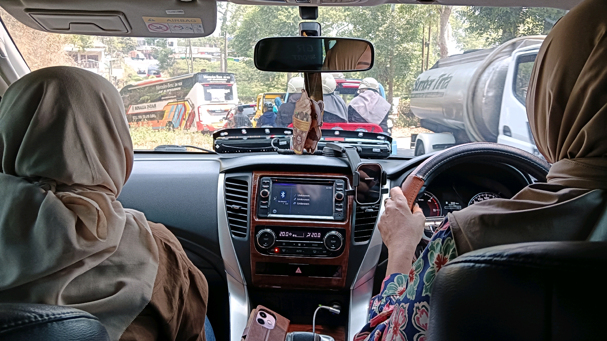 The image, taken inside a car, shows a view from the passenger seat looking towards the driver and the road ahead. The driver, wearing a beige hijab and a floral-patterned shirt, is seen from behind, with their hands on the steering wheel. The car's interior features a dashboard with a media system, climate controls, and a phone mount. Outside the car, traffic is visible, including buses and a large tanker truck. The background suggests a semi-urban or rural setting with trees and buildings lining the road. The presence of traffic and the surrounding environment indicates the car is likely on a journey. The location is identified as Ngargoyoso, Indonesia.