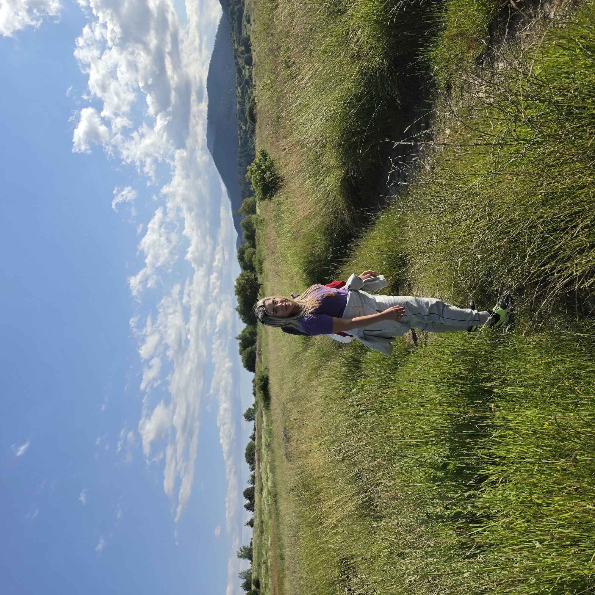 A young woman stands on a dirt path in a grassy field under a blue sky with scattered clouds. She is wearing a purple t-shirt, grey pants, and athletic shoes. A grey jacket is tied around her waist. In the background, there are trees and a distant mountain range. The scene appears to be in or near Ravnogor, Bulgaria, a location known for its natural beauty and hiking trails.
