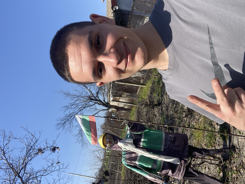 A young male, with short dark hair and a grey Nike t-shirt, is shown in a frontal selfie. He is smiling and displaying a two-finger peace gesture with his right hand. The scene is an outdoor setting, identified as a garden or cultivated area within the city of Merichleri, Bulgaria. Behind the male, a scarecrow is positioned to the left, dressed in a yellow hard hat, a multi-paneled jacket featuring green, grey, and dark blue/purple sections with reflective elements, a white scarf, and dark trousers. A Bulgarian flag, horizontally striped in white, green, and red, is visible on a pole further back, to the right of the scarecrow. The environment consists of disturbed soil with sparse green growth, several bare deciduous trees, and rudimentary structures with fencing, all under a clear blue sky.