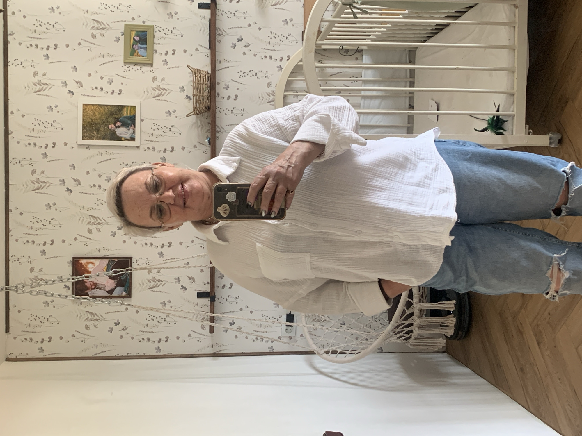 An adult woman wearing glasses, a white button-down shirt, and blue jeans stands indoors, holding a smartphone. The phone case is dark with light-colored embellishments. She appears to be taking a selfie. The background includes wallpaper with a floral pattern, three framed pictures, a wicker basket attached to the wall, a white baby crib, and a hanging macrame chair. The floor is wooden. The scene is located in Chaikovsky, Russia.