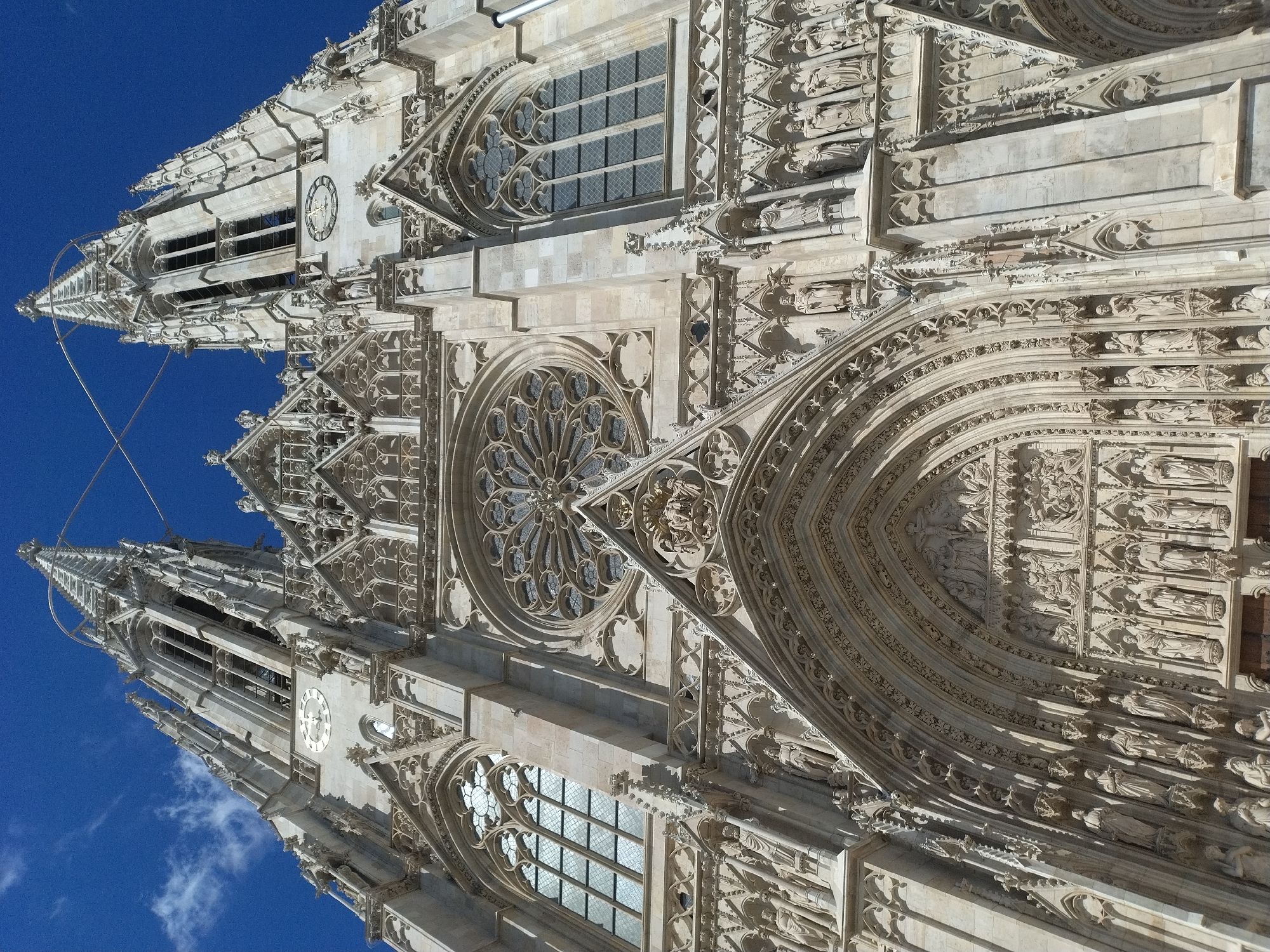 The image shows a low-angle, full shot of the facade of St. Stephen's Cathedral in Vienna, Austria. The cathedral, constructed from light-colored stone, is highly ornate with Gothic architectural elements. The sky above is a vibrant, clear blue with a few wispy white clouds.
Dominating the central portion of the facade is a large rose window, intricately carved. Below the rose window, a grand arched entrance is visible, adorned with numerous detailed sculptures of figures. Flanking the central rose window are tall, pointed spires characteristic of Gothic cathedrals, each featuring arched windows and decorative stonework. A clock face is visible on the right spire. The facade is further embellished with a multitude of sculptures, niches, and complex tracery. The lower section of the facade includes multiple levels of arched doorways and windows, all richly decorated. The overall impression is one of grandeur and intricate craftsmanship.
