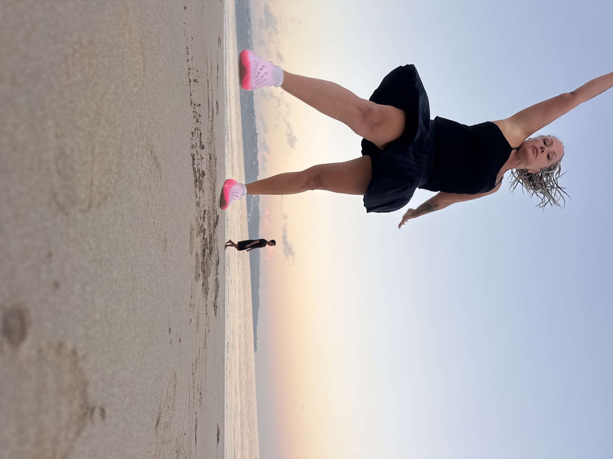 The image depicts a woman in mid-air, jumping on a sandy beach at sunset. She is wearing a black tank top and a black skirt, with bright pink shoes. Her hair, styled in thin braids, is spread out behind her. Her left arm is raised upwards, and her right arm is extended outwards. In the background, a man is walking on the beach near the water. The sky is a soft gradient of blue and pink, indicative of a sunset. The location is identified as Kuta, Indonesia, with the ocean visible and a distant shoreline. The perspective is low, looking up at the jumping woman, emphasizing her movement. The sand in the foreground is detailed, showing footprints and textures.