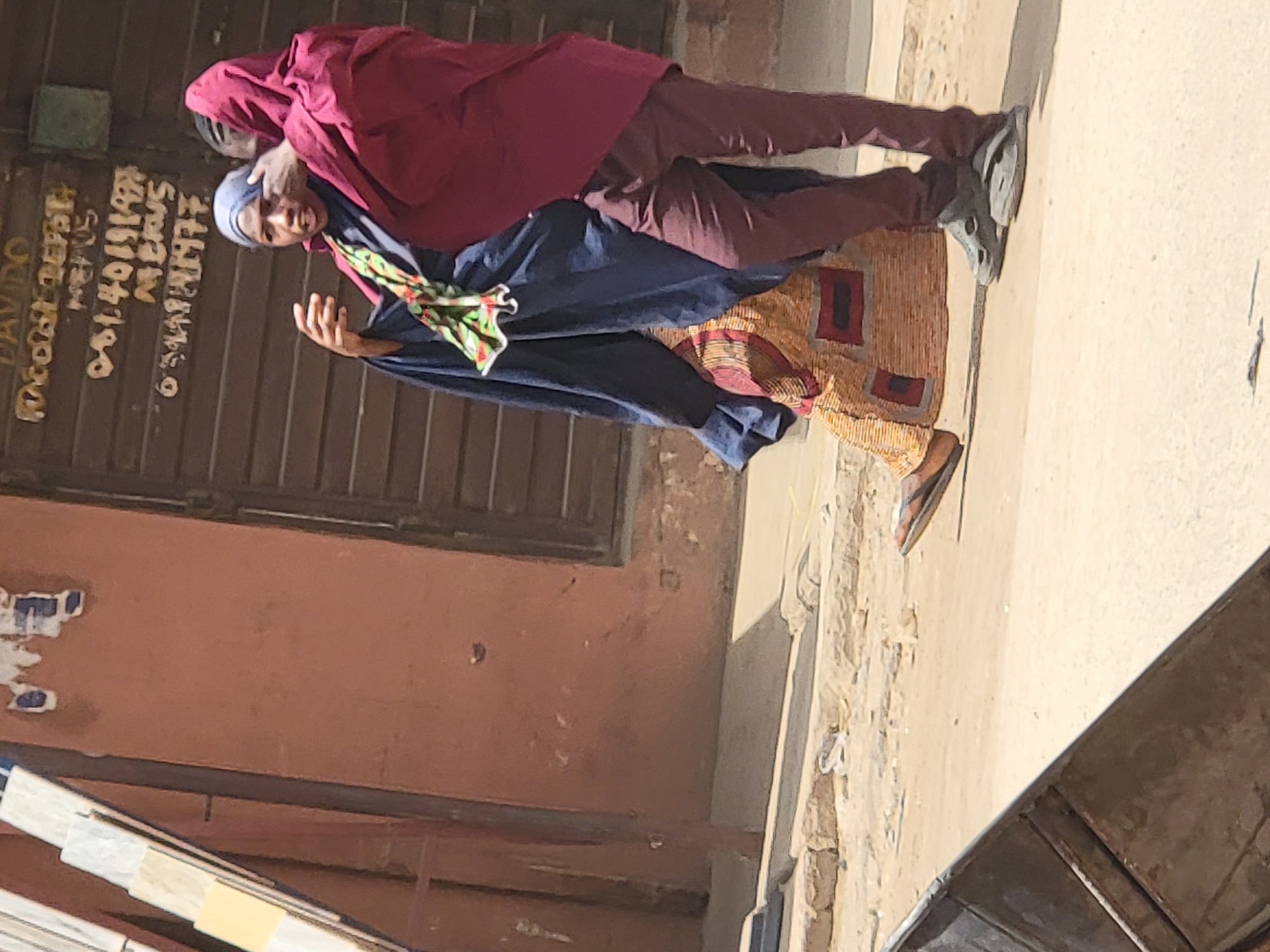 On a street in Maiburiji, Nigeria, two people stand on a sidewalk next to a building with a closed, metal shutter door on which text and numbers are painted. The person closest to the building wears a maroon head covering, maroon trousers, and gray shoes, and has a hand placed on the head of the other person. The other person wears a blue head covering, a dark blue garment, a patterned yellow and brown dress with red squares, and brown sandals. This person has one hand raised in the air. The building is peach-colored with patches of peeling paint. The sidewalk is light brown. A gray metal structure is visible at the bottom right of the frame, and objects that appear to be building materials are visible at the bottom left.