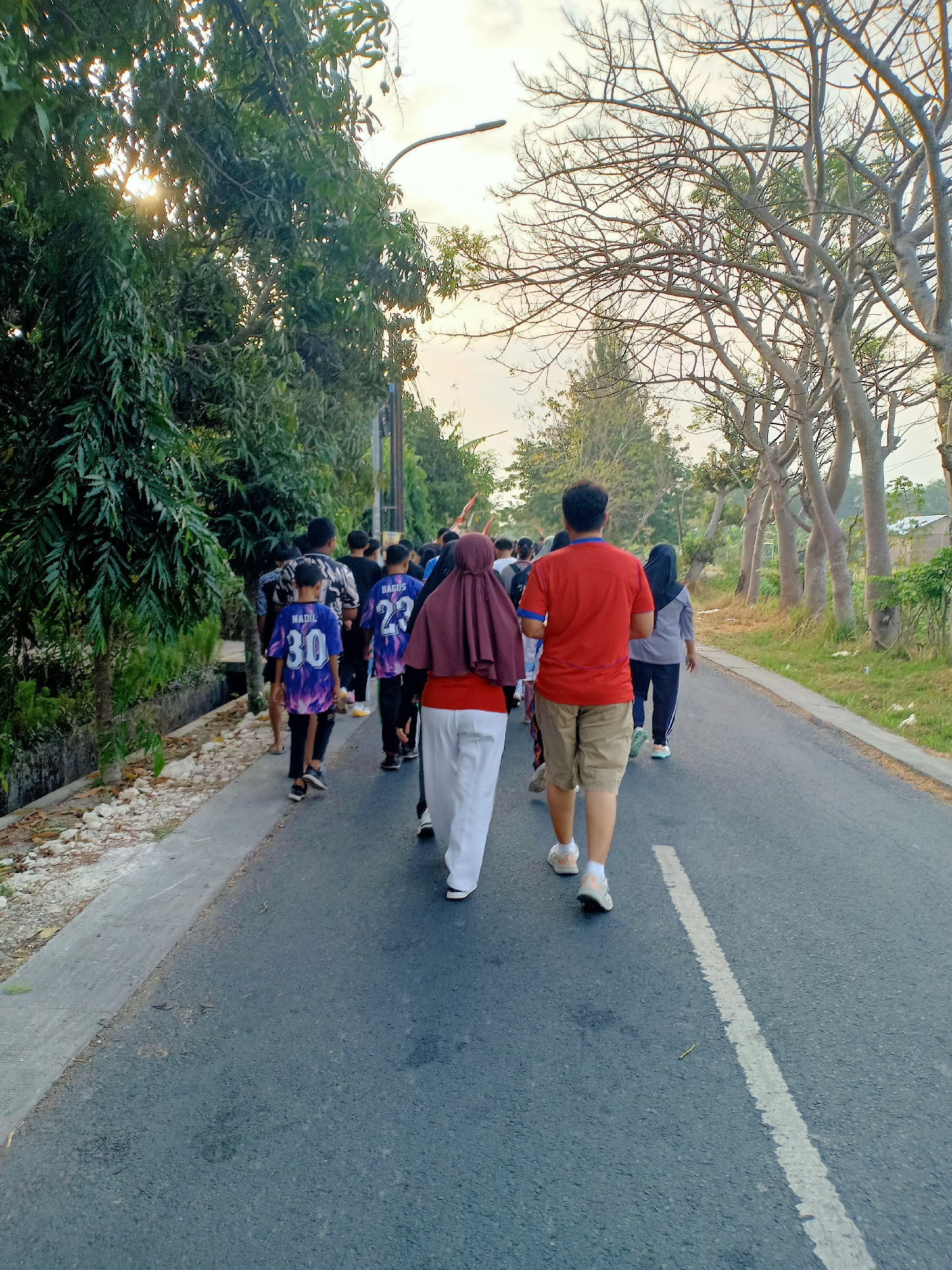 In the city of Mojoranu, Indonesia, a group of people walks along a paved road. The individuals are moving away from the viewer. The road has a white dashed line painted down the center. To the left of the road is a raised concrete sidewalk with rocks and vegetation. Further left, lush green trees line the area. To the right of the road, bare trees with thin branches are visible, along with grass and some residential structures in the background. The sky is overcast with a pale, diffused light, suggesting it is either early morning or late afternoon. The people are dressed in casual clothing, including t-shirts, pants, and some are wearing headscarves. Some individuals are wearing sports jerseys with numbers. The overall impression is of a group engaged in a procession or an organized walk.
