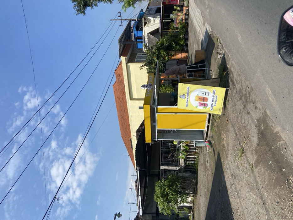 A street scene in Mojosari, Indonesia, located at coordinates -7.503 latitude and 112.54 longitude. A small yellow kiosk with a sign that reads "Teh Point Indonesia" and "Harga Mulai 3000" is situated on the right side of the street. A woman in a headscarf stands at the kiosk, seemingly tending to the business. There are multiple power lines running overhead against a bright blue sky with scattered white clouds. Several buildings line the street, some with terracotta tile roofs and others with corrugated metal roofs. Green foliage, including trees and bushes, are present on the sides of the road. The street itself is paved with gray asphalt and lined with dirt shoulders. There is a motorcycle parked in the foreground, partially obscuring the view. The overall environment is typical of a residential area in a smaller Indonesian town.