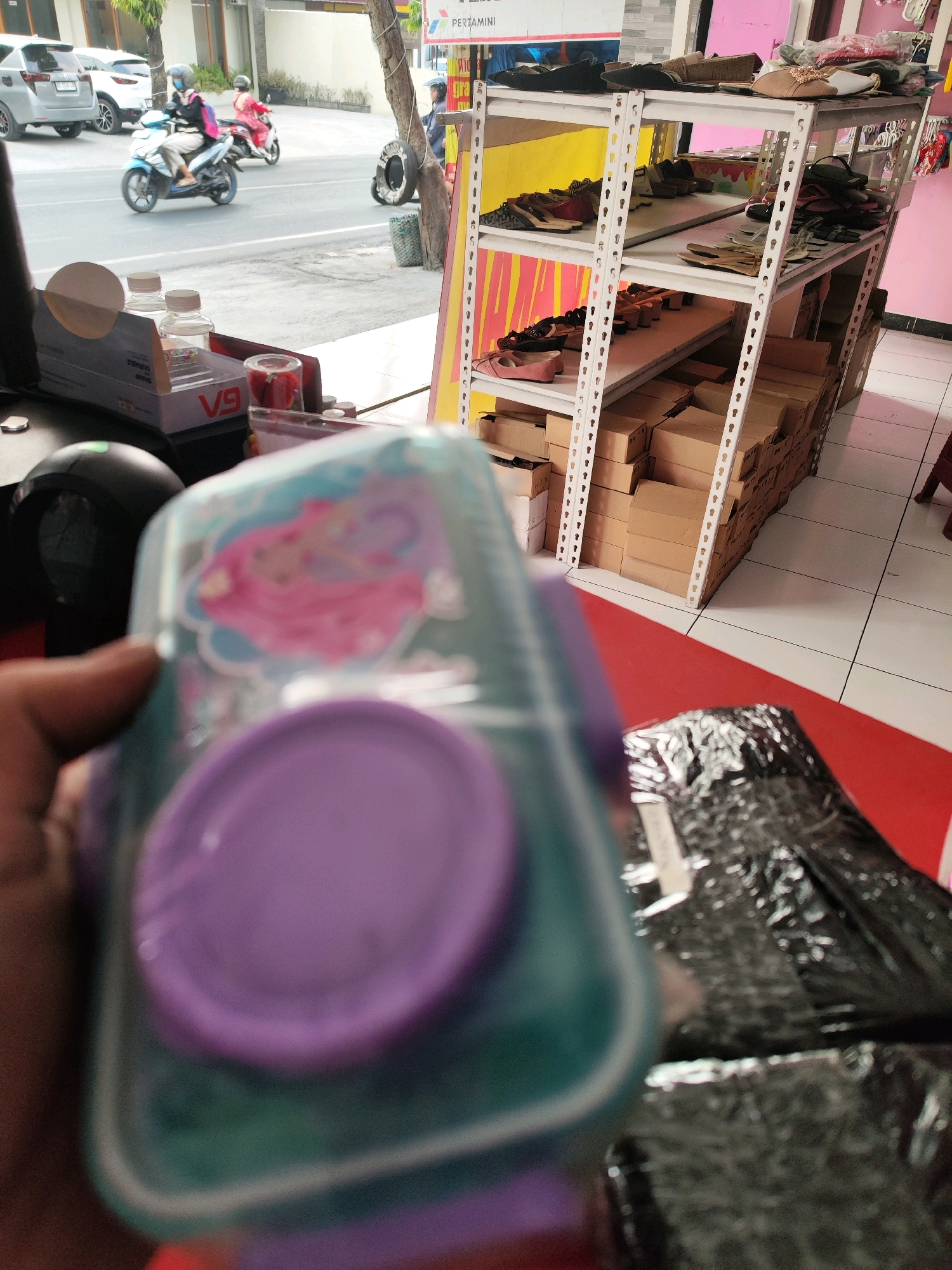 The image depicts a street scene in Sragen, Indonesia, with a focus on a display shelf of shoes and boxes. In the foreground, a person's hand is visible, holding a transparent plastic container with a purple lid, featuring a cartoon character. Behind this container, various products are displayed on a white metal shelf unit. The shelf holds numerous pairs of shoes, including sandals and closed-toe footwear, arranged on different levels. Below the shelves, stacks of cardboard boxes are neatly piled. The background shows a street with vehicles, including cars and motorcycles, and a sidewalk. The environment appears to be an outdoor or semi-outdoor commercial area.