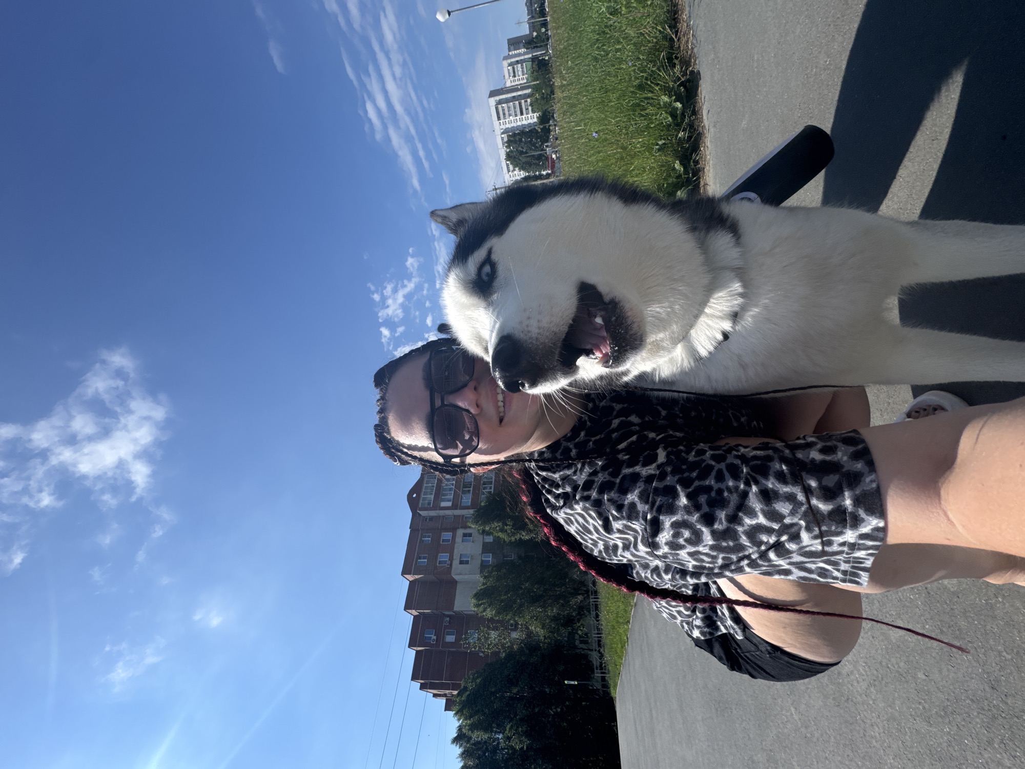 In a selfie taken outdoors, a young woman with dark braided hair and sunglasses smiles at the camera, crouching beside a happy-looking husky dog. The woman wears a black and white leopard-print top and black shorts. The husky has striking blue eyes and its mouth is open as if panting. Behind them, a paved path runs alongside a grassy area, with buildings and trees visible in the background under a bright blue sky with scattered clouds.