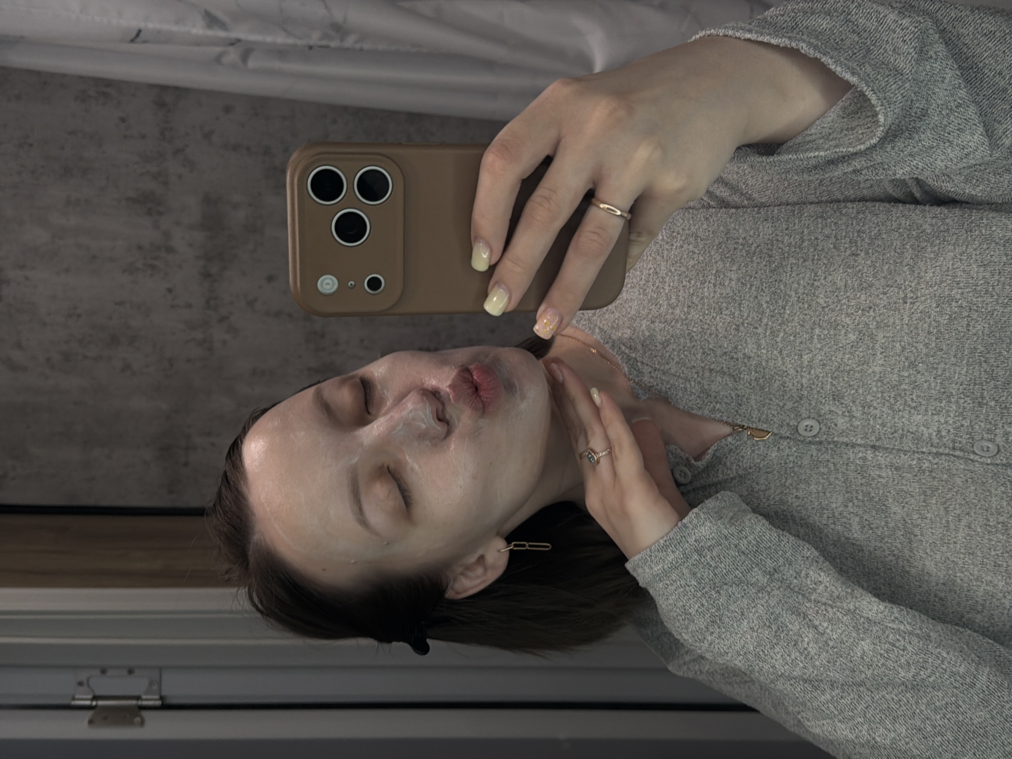 An indoor selfie, captured at a 90-degree clockwise rotation, depicts a person with a white facial mask applied to their face. The individual holds a tan-colored smartphone with a prominent triple-lens camera module, a flash, and other small sensors, consistent with a modern smartphone model like an iPhone. Their eyes are closed, and lips are gently pursed in a slight pout.
The person is wearing a light grey, possibly knitted or textured, long-sleeved top or cardigan with buttons. They have rings on their fingers: a simple gold band on the index finger of the hand holding the phone, and another ring with a darker stone on the ring finger of the other hand, which is resting on their cheek. Their fingernails are painted with a soft yellow or cream-colored polish, with one nail featuring a subtle sparkly or textured design. A gold-colored earring, resembling a paperclip chain, hangs from their left ear. Dark hair frames their face, tucked behind the ears.
The background is an indistinct indoor setting. A grey, textured wall or surface is visible behind the person, and a lighter ceiling or wall area can be seen in the upper part of the frame. Hints of a window frame or dark furniture are visible towards the bottom of the image. The scene suggests a moment of personal care and relaxation, likely captured for social sharing. The location context provided is Reshetnikovo, Russia. There is no visible text in the image.
