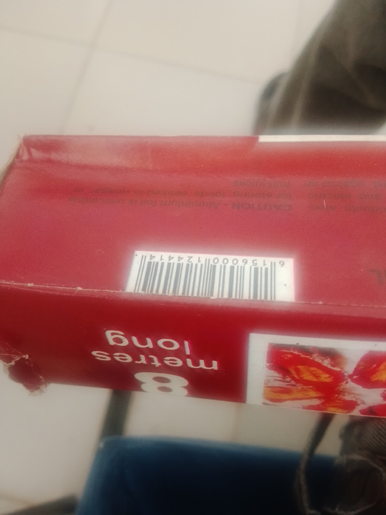 The image shows a close-up of a red cardboard box, likely containing kitchen foil or similar wrap. White text on the box reads "8 metres long" and "CAUTION - Aluminium foil is unsuitable". There is also a barcode with the number "6 156000 124414". The box is slightly damaged on the left side, with a tear in the material. A small, colorful image is visible on the right side of the box. The background is blurred, suggesting the photo was taken indoors. The scene is set in an unspecified urban environment.