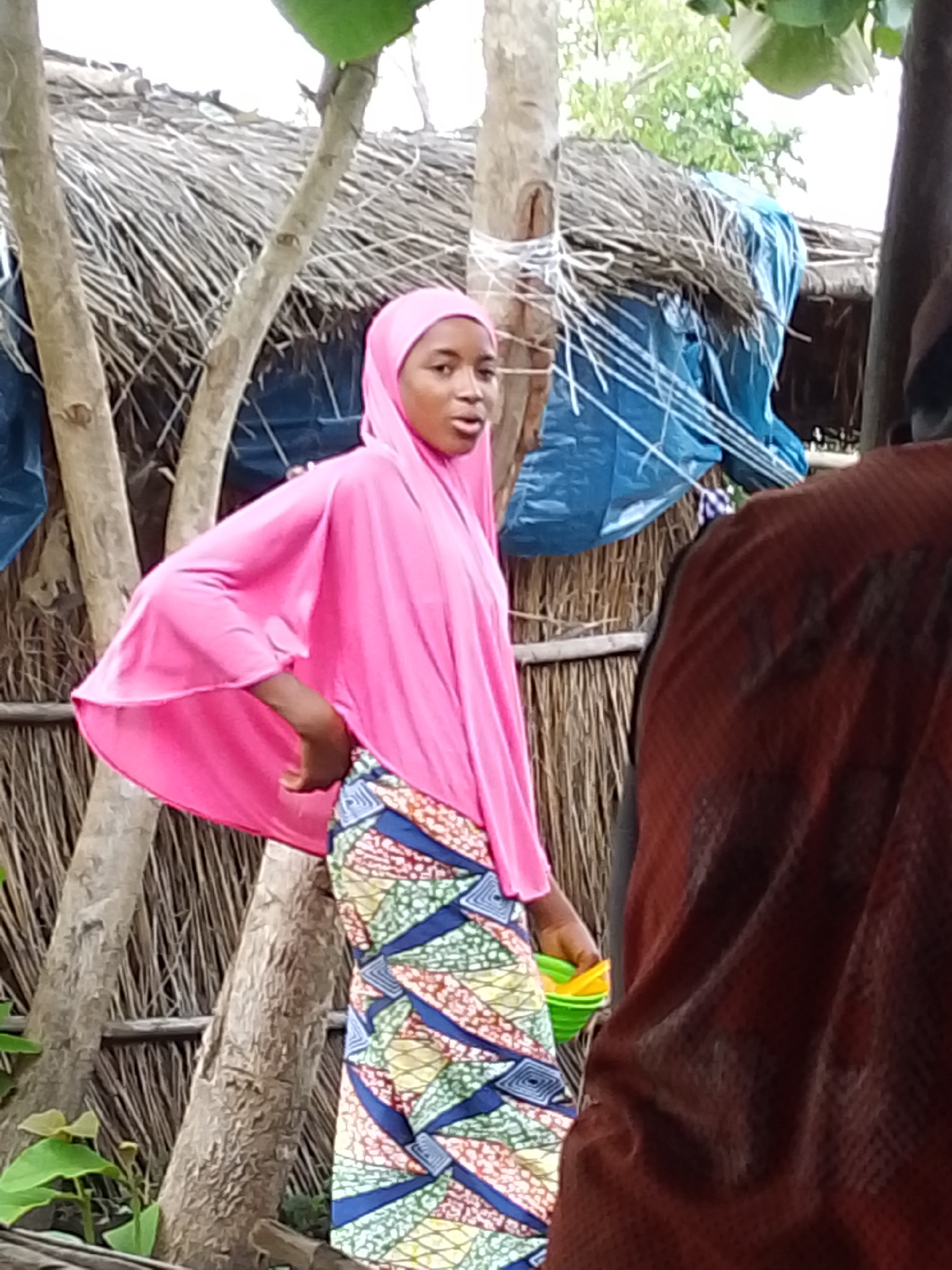 In Otulu, Nigeria, a young girl wearing a bright pink hijab and a patterned skirt stands near a thatched hut. She is looking towards the camera with her mouth slightly open, possibly mid-sentence or surprised. To her right, a person in a dark reddish-brown garment is partially visible, obscuring the view of the hut's interior. The hut's roof is made of dried thatch, and a blue tarp is draped over one side, held in place by white twine. The scene is set against a backdrop of trees and foliage, suggesting an outdoor, possibly rural, environment. The girl is holding a small, multi-colored container in her left hand.