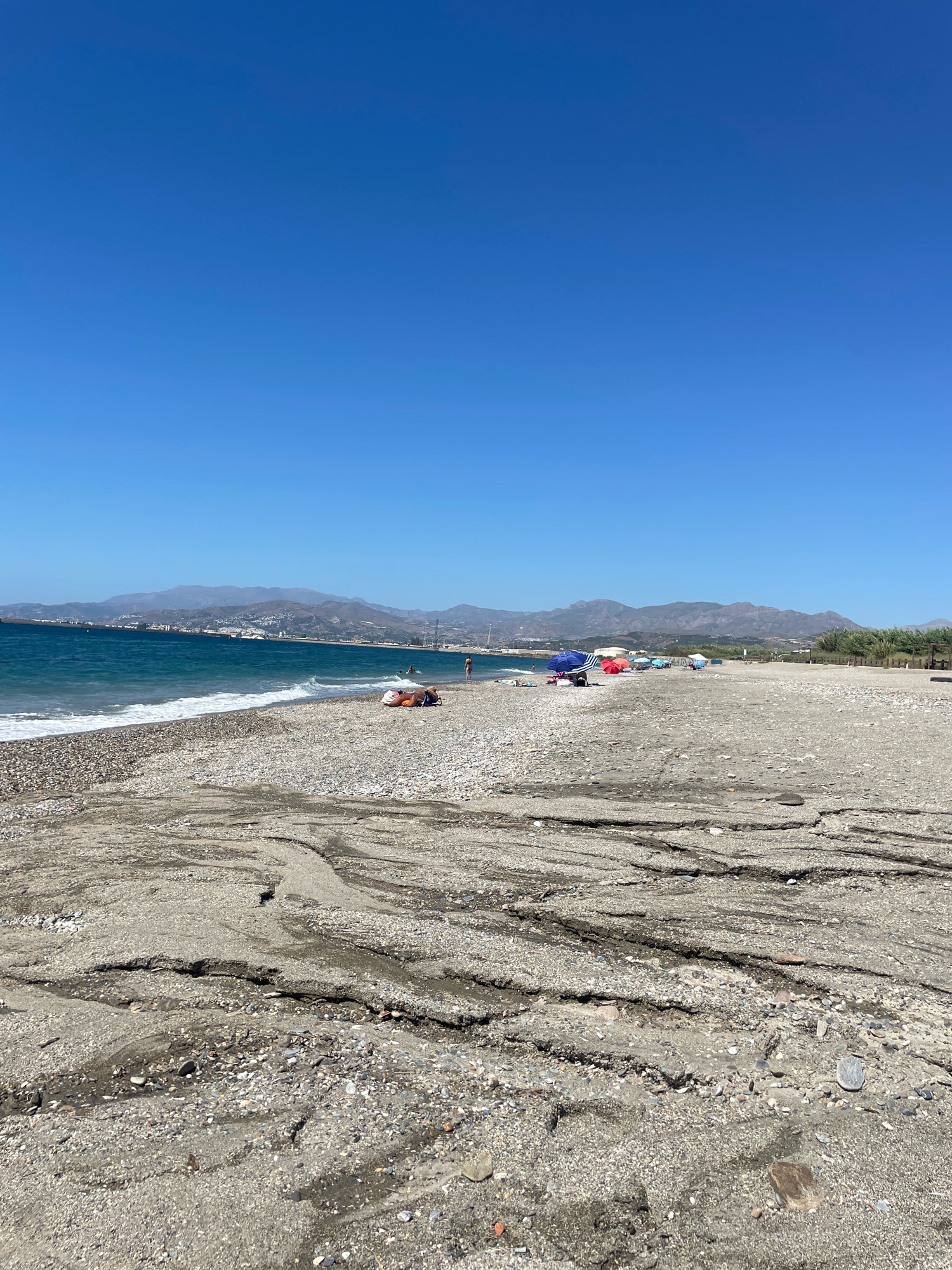 A wide shot of a beach in Torrenueva Costa, Spain, on a clear, sunny day. The foreground is dominated by the pebbly and sandy texture of the beach, showing channels carved by water. In the mid-ground, the blue sea with gentle waves meets the shore. Along the waterline, several beach umbrellas and chairs are visible, with a few people scattered on the sand. Further back, a coastal town with buildings is nestled against rolling hills that form the backdrop. The sky is a clear, bright blue.