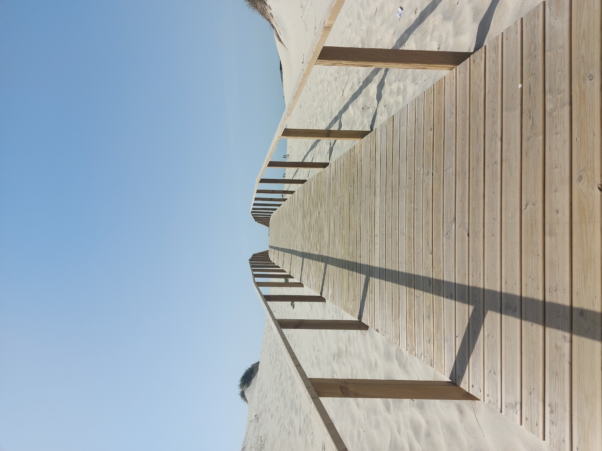 The image shows a wooden boardwalk in Gafanha da Nazaré, Portugal, leading through sand dunes under a clear blue sky. The boardwalk has a simple wooden railing on both sides, and the shadows of the railing posts fall diagonally across the wooden planks. The sand dunes appear light in color and are sparsely vegetated.